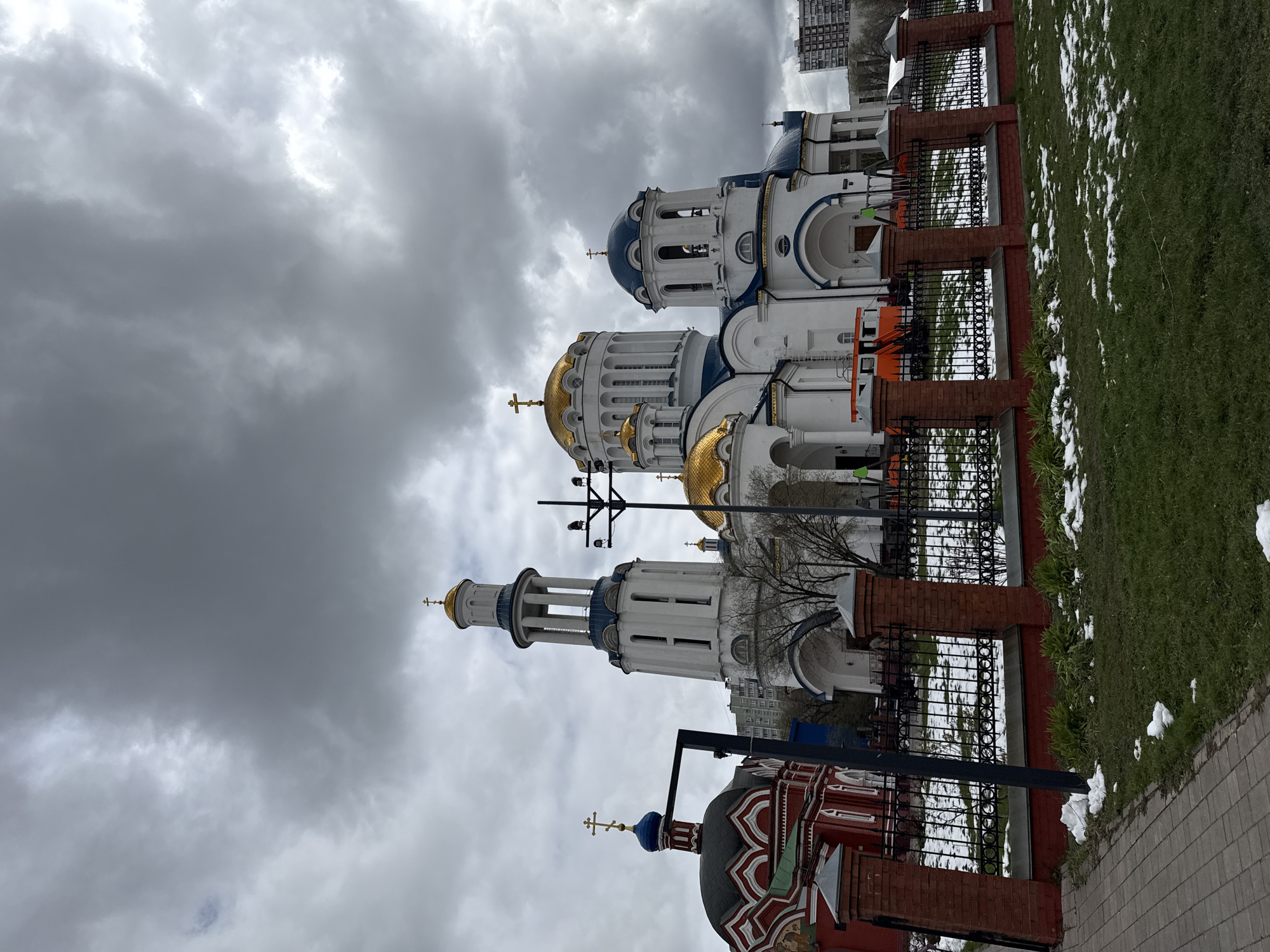 The image displays a complex of Orthodox Christian churches under a heavily overcast, gray sky. On the left, a large white church with multiple blue and golden onion domes is prominent, featuring several bell towers topped with crosses. To its right, and slightly in the foreground, another smaller church is visible, constructed of red brick with patterned, darker domes, one of which is blue.
A black metal fence with brick pillars runs horizontally across the middle ground, separating the church complex from a grassy area on the right. This grass is interspersed with patches of melting snow, indicating a cool season, likely late winter or early spring. A paved sidewalk borders the bottom edge of the image. In the background, behind the churches and bare trees, a tall, multi-story building can be seen.
The weather is characterized by thick, dark clouds, giving the scene a somber or subdued atmosphere during what is clearly daytime. No people or specific activities are depicted; the scene is static, focusing on the architectural structures. The distinct architectural style, including the onion domes and Orthodox crosses, strongly suggests a Russian setting, consistent with Moscow. No visible text is legible in the image.