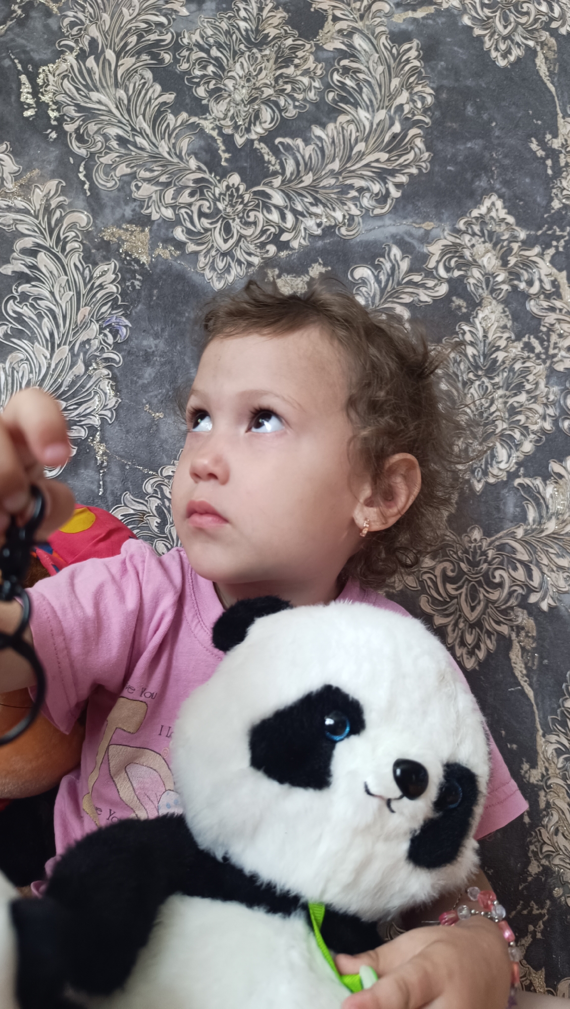 A young girl with curly brown hair is looking upwards, holding a plush panda toy. She is wearing a pink t-shirt. The background is a patterned wallpaper with floral motifs in shades of gray and gold. The image is a close-up shot, focusing on the girl and the toy. There is no indication of the city of Karsha, Russia, in the image.