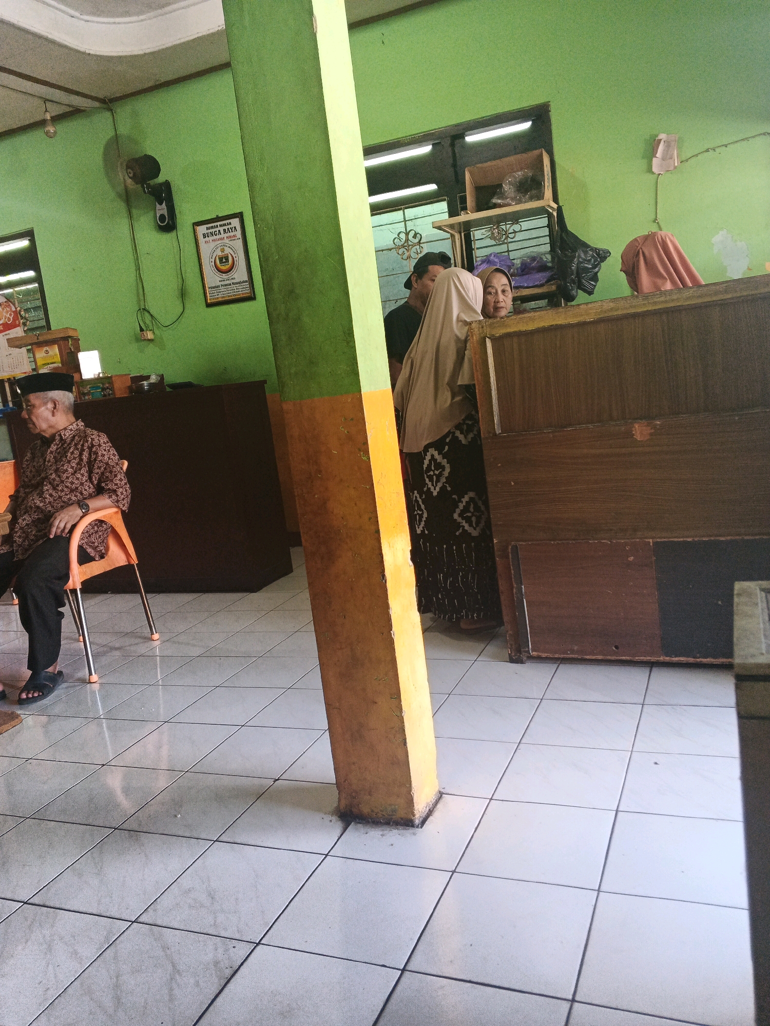 In Ungaran, Indonesia, inside a simple restaurant with light green walls, a man in a patterned brown shirt and black pants sits on an orange chair. He is wearing black slippers and a black cap. To his right, a thick, yellow and green painted pillar stands in the foreground. Behind the pillar, two women are partially visible. One woman, wearing a light brown hijab and a patterned skirt, stands looking towards the left. Another woman, wearing a dark hijab, stands next to her. Wooden counters and shelves are visible in the background, containing various items. A fan is mounted on the wall to the left, and a framed certificate hangs nearby. The floor is tiled with white squares. The scene suggests a casual dining environment.