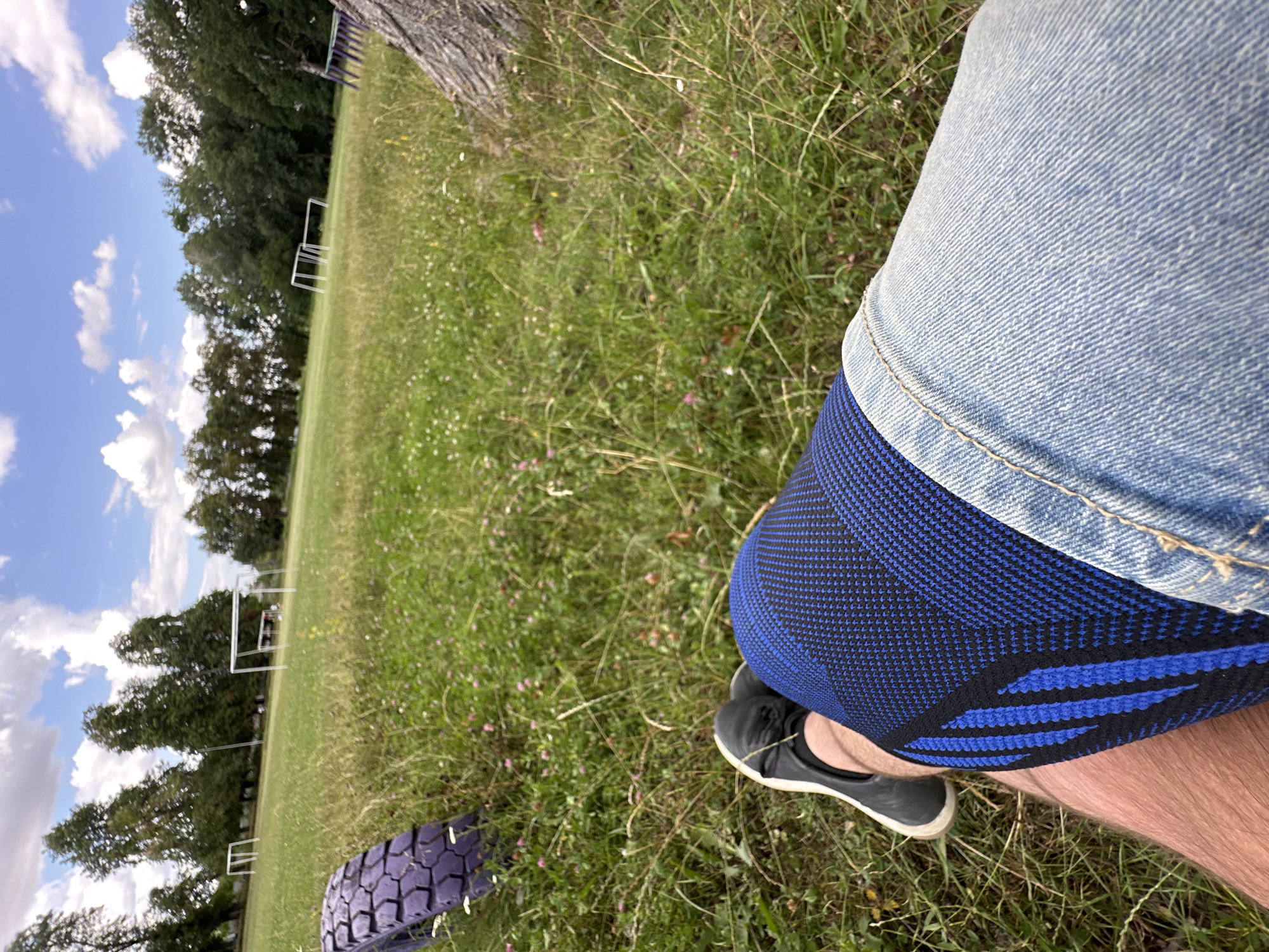 A person is sitting on a grassy field. The person is wearing blue and black patterned compression sleeves on their legs, and blue denim shorts. A black sneaker is visible on their left foot. In the background, there are two soccer goals and a line of trees. To the left of the frame, a purple tire lies on the grass. The sky is partly cloudy. The location is a soccer field in the village of Makiv, Ukraine.