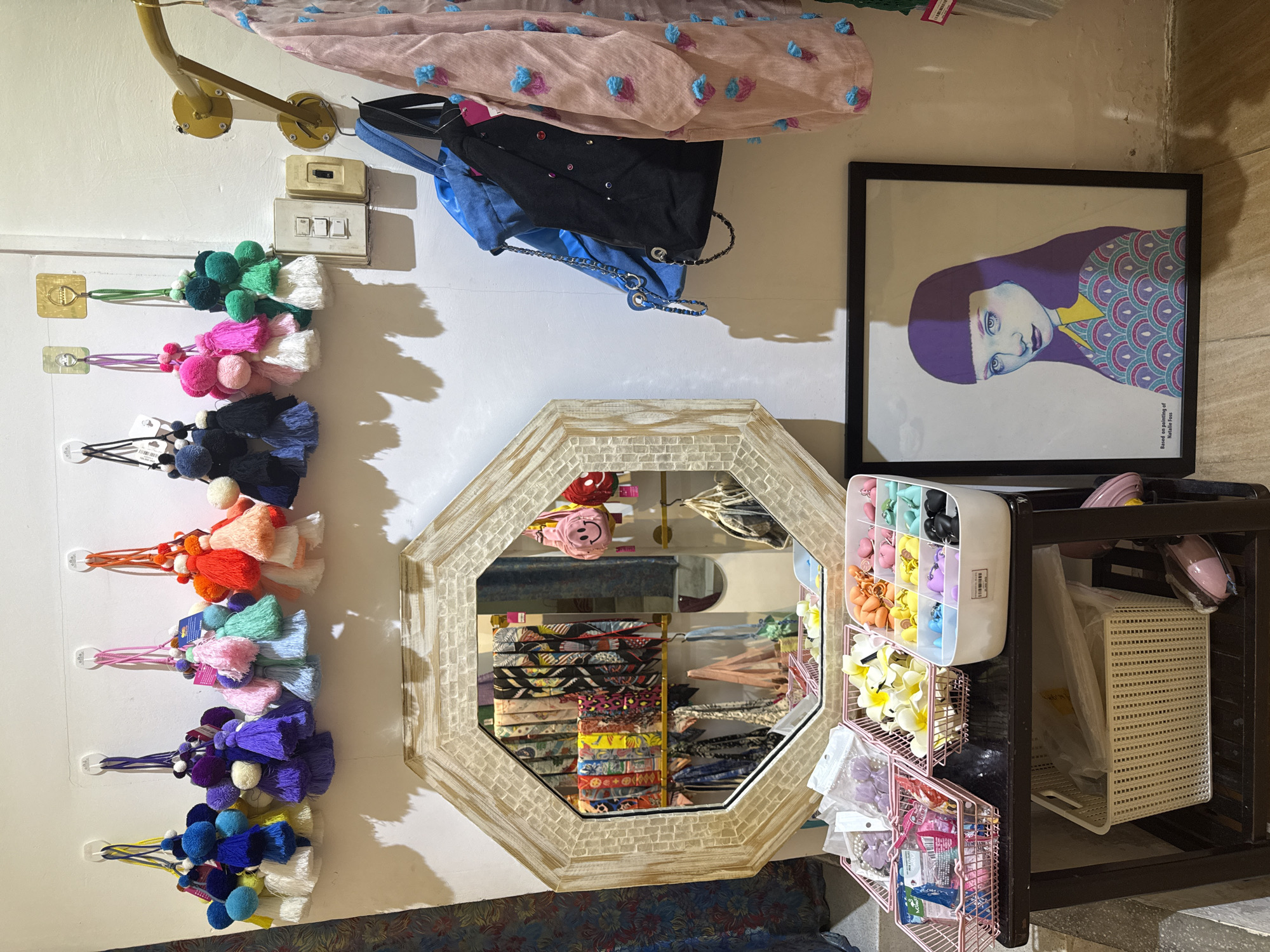This is a medium shot, eye-level, indoor photograph taken in a retail establishment. The scene depicts a display of various items, including decorative tassels, handbags, clothing, a mirror, storage baskets filled with small accessories, and a framed art piece.
In the upper portion of the image, a row of colorful pom-pom and tassel keychains hangs from white wall hooks. These keychains are in various colors such as blue, purple, pink, orange, and green.
To the right of the keychains, a gold-colored metal rod extends horizontally, supporting a light pink, sheer garment adorned with blue pom-poms. Below this rod, a blue canvas tote bag and a black tote bag with chains and embellishments are hung.
A large, octagonal mirror with a textured, light-colored frame is positioned centrally in the image. The mirror reflects a display of colorful scarves arranged on a rack behind it. Small pink emoji-shaped keychains are also visible hanging on the right side of the mirror.
In the foreground, a dark wooden table holds several pink wire baskets and a white plastic organizer. The baskets contain various small items like charms, beads, and hair accessories in shades of purple, yellow, orange, and blue. A larger white woven basket filled with plastic-wrapped items sits below the table.
To the right of the table, a framed artwork featuring a stylized portrait of a woman with purple hair and a colorful patterned shirt leans against the wall. The text at the bottom of the frame indicates it is "Based on painting of Natalie Foss."
The background wall is white. The floor appears to be tiled. The overall impression is a retail display likely selling accessories and clothing. The establishment is located in Nha Trang Ward, Vietnam.