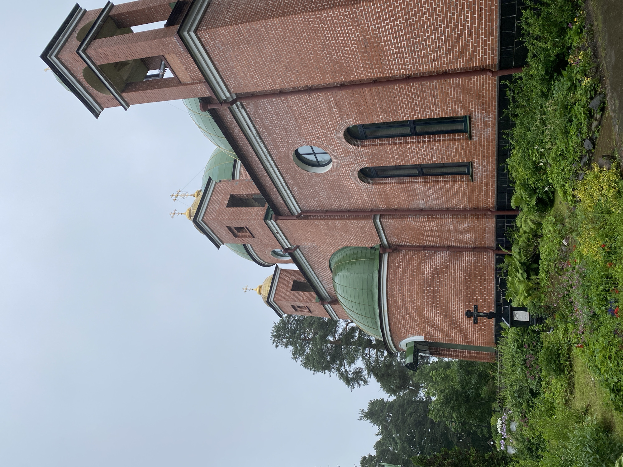 The image depicts the exterior of the Roman Catholic Church of the Sacred Heart of Jesus, located in Priozersk, Russia. The church is constructed of red brick and features several green domes topped with golden crosses, as well as a bell tower. The surrounding area is lush with greenery, including trees and various plants. In the foreground, a black cross with a crucifix stands amidst the foliage, and beyond it, there appear to be some grave markers, suggesting a cemetery or memorial garden adjacent to the church. The sky is overcast, indicating a cloudy day.