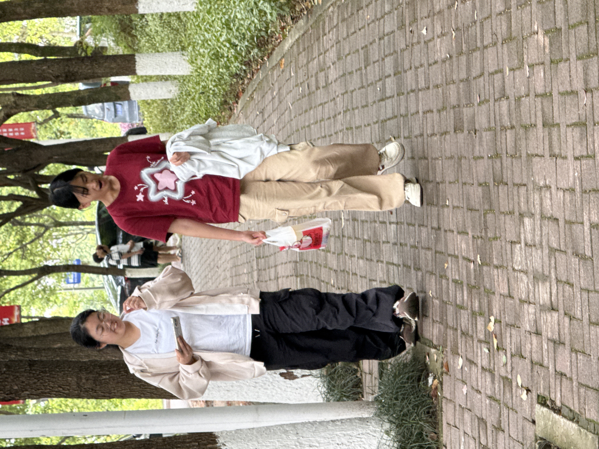 Two women are walking along a brick-paved sidewalk adjacent to a street lined with trees.
The woman on the right wears a burgundy shirt with a star design, beige pants, and white sneakers. She carries a white cloth bundle and a small red and white bag.
The woman on the left wears a white t-shirt, black pants, a beige jacket, and gold sneakers. She is holding a phone in her hand.
The location appears to be Jiangjin, China.