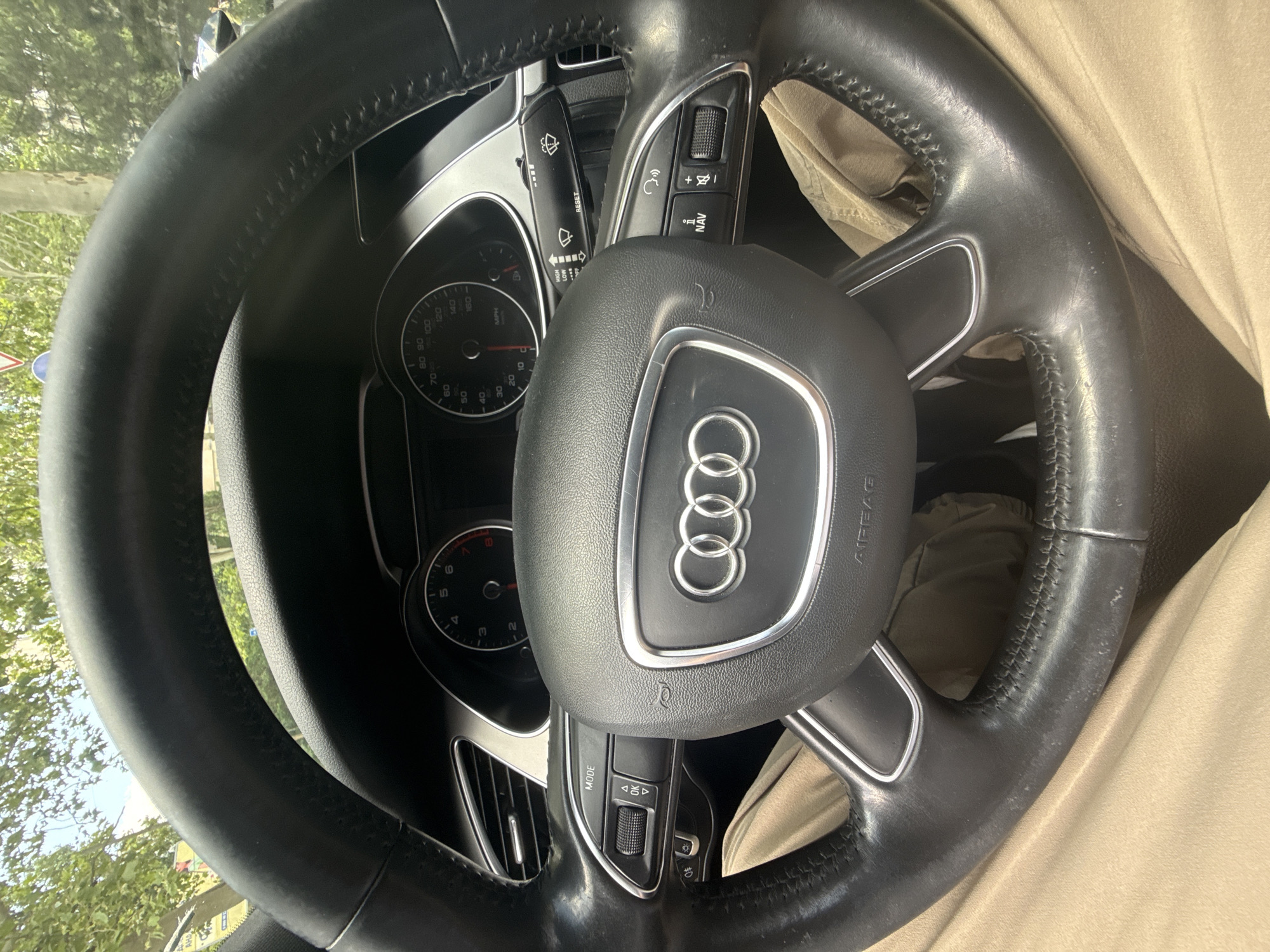 A first-person perspective shot shows the interior of an Audi vehicle from the driver's seat. The leather-wrapped steering wheel, emblazoned with the Audi rings, is central to the frame. On the left side of the steering wheel, a "MODE" button is visible, along with a dial and up/down arrow buttons. On the right side, a stalk with "HIGH," "LOW," and a windshield wiper icon is present, along with a "RESET" button. Further right, there's a stalk with icons for a phone and volume control, alongside "NAV" and "+" and "-" buttons. The dashboard includes a speedometer and tachometer, with the speedometer showing a maximum of 180 MPH. The driver's legs, clad in tan trousers, are visible at the bottom of the image. Outside the car, through the windshield, green trees and a blue sky can be seen, suggesting an outdoor setting, possibly in a city given the presence of traffic signs.