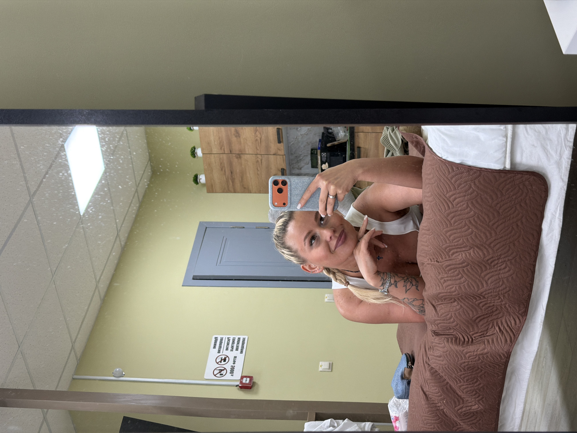 An indoor mirror selfie captures a woman with blonde hair, styled in two braids, wearing a white top. She is smiling softly and holding a smartphone with a light grey and orange case, revealing a ring on her finger. Tattoos are visible on her left arm, and she wears a small cross necklace.
The reflection in the mirror shows a room with light yellow-green walls and a light-colored acoustic tile ceiling featuring a prominent, bright, diamond-shaped light fixture. In the background, there is a dark grey door and a cabinet with wooden doors, on top of which small potted plants are arranged. A small, white, dome-shaped object is mounted on the wall, possibly a camera or sensor.
In the foreground of the reflection, the woman is partially resting on a brown quilted pillow or blanket, which lies on white bedding or cushions. A small blue bag or purse is also visible near the bedding.
On the wall, reflected and thus appearing reversed, is a white sign with black and red symbols and Cyrillic text. The symbols include a crossed-out cigarette and a finger to lips indicating quiet. The text, when un-reversed, reads "КУРИТЬ ЗАПРЕЩЕНО" (Smoking Forbidden) and "ШУМЕТЬ ЗАПРЕЩЕНО" (Noise Forbidden), and possibly "С 20:00" (From 8:00 PM), suggesting quiet hours. Below the sign is a red fire alarm pull station. The scene appears to be a public or commercial interior, possibly a waiting room or treatment facility.
