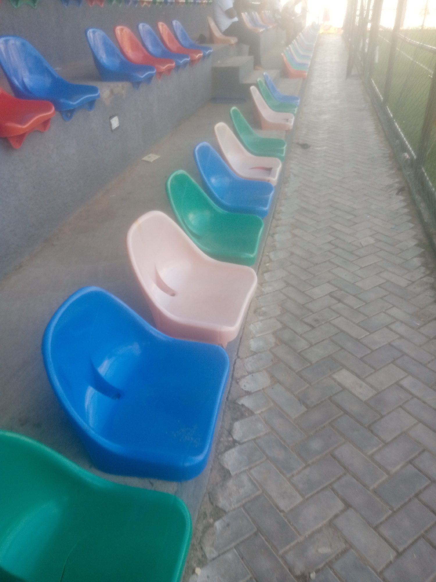 The image depicts a row of empty, colorful plastic stadium seats arranged on concrete tiers. The seats are primarily blue, red, and a light peachy color, with a few green ones visible in the foreground. The stadium appears to be outdoors, with a paved walkway made of interlocking gray bricks running alongside the seats. In the background, a chain-link fence and some greenery are partially visible. The lighting suggests it is daytime, possibly late afternoon or early morning, due to the soft, diffused light. There are no people directly interacting with the seats in the frame. The context of Jalingo, Nigeria, is provided, suggesting this is likely an outdoor sports or event venue within the city.