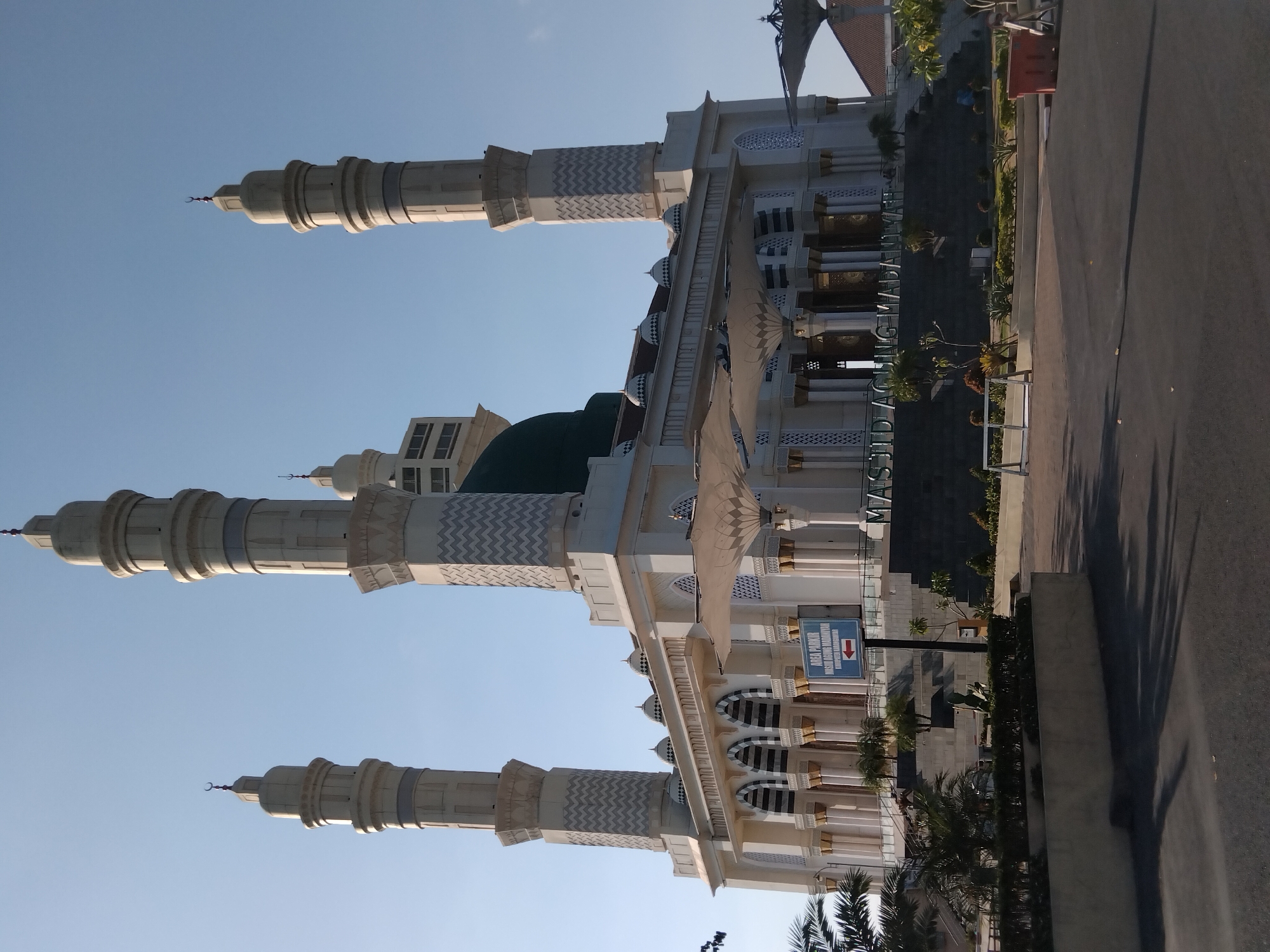 The image depicts the Masjid Agung Madaniyah in Karanganyar, Indonesia, on a clear, sunny day. The mosque is a large, white building with a prominent green dome and four tall, slender minarets. The minarets are adorned with intricate patterns and topped with crescent moons. A sign near the entrance reads "AREA PARKIR MASJID AGUNG MADANIYAH KABUPATEN KARANGANYAR" with an arrow pointing left. The mosque is surrounded by a paved area, some landscaping with trees and shrubs, and a series of steps leading up to the main entrance. The overall scene conveys a sense of grandeur and tranquility.