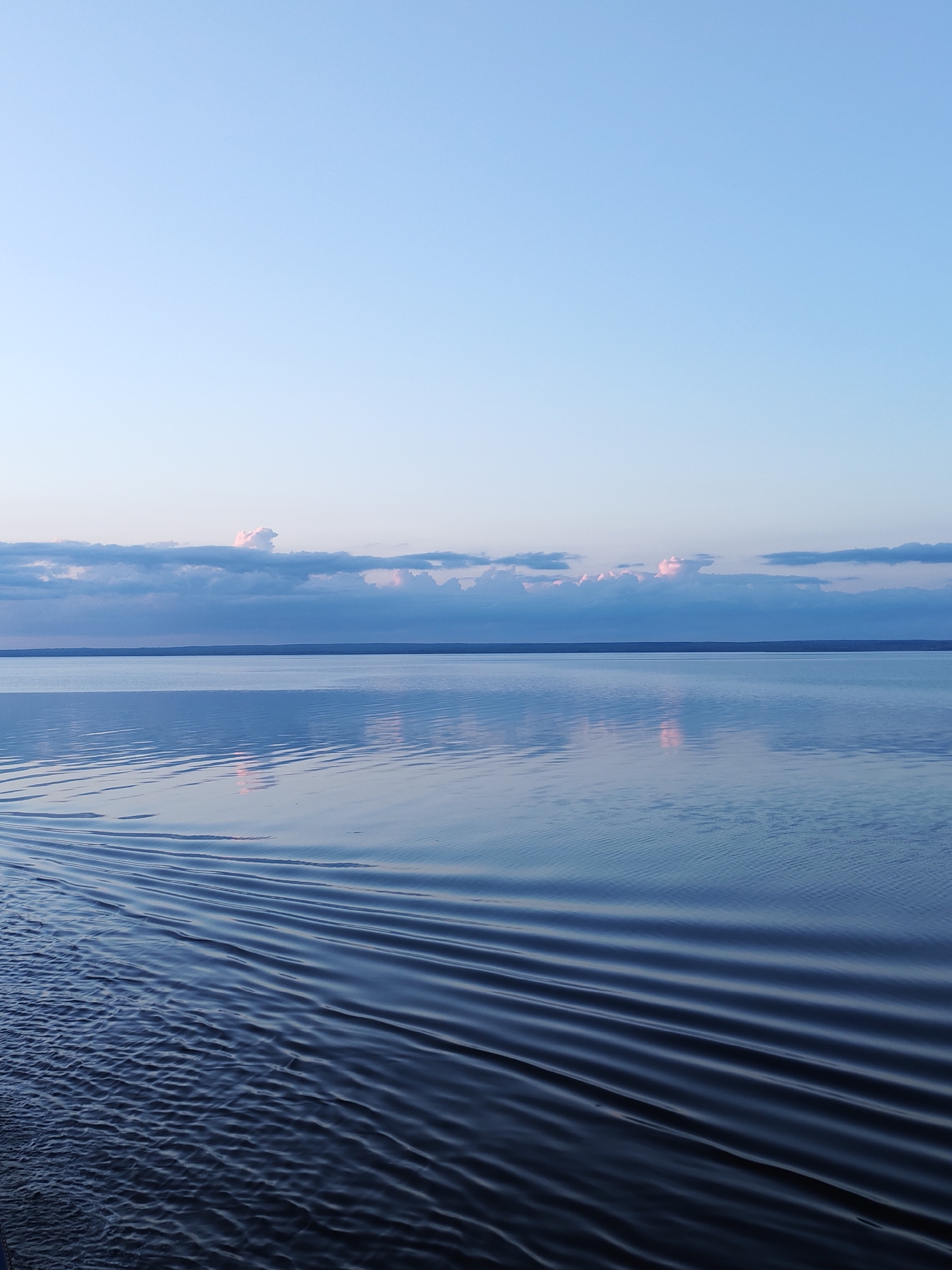 The image depicts a body of water, likely a lake or a sea, under a clear, light blue sky transitioning to slightly darker shades near the horizon. A thin line of land is visible in the distance. The water's surface is partially rippled, with darker, more defined waves in the foreground and smoother reflections in the midground. The scene appears tranquil and natural. The location is unspecified.