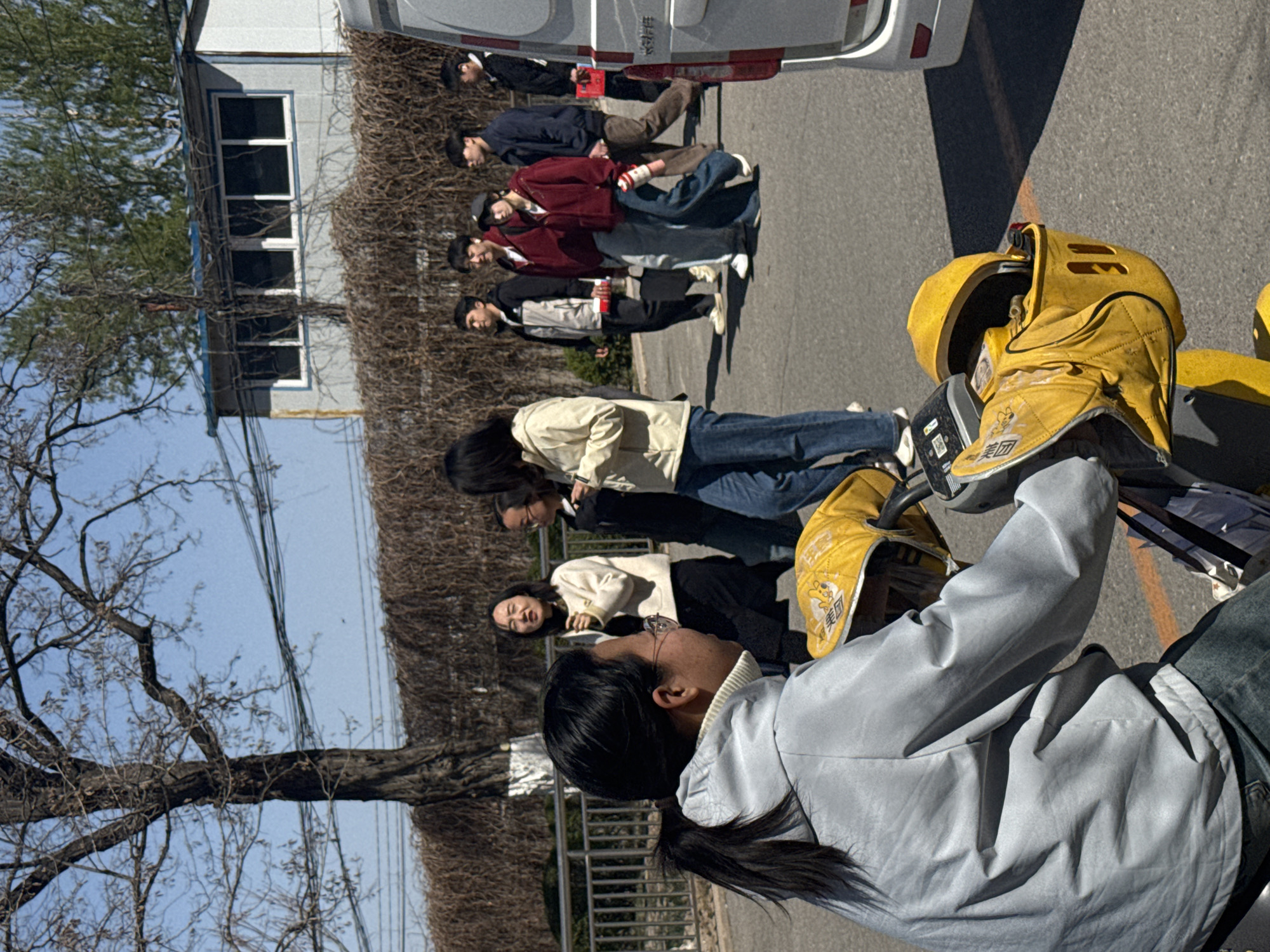 The image depicts an outdoor scene in Zhongtun, China, under bright daylight and a clear blue sky, suggesting a sunny day in late winter or early spring due to the bare trees and vines.
In the foreground, a person is seen from behind, wearing a light grey jacket and jeans, actively engaged with bright yellow delivery equipment. This equipment, including a helmet and possibly a bag, prominently displays the Chinese characters "美团" (Meituan) and features a cartoon mascot, indicating its association with the Meituan food delivery service.
In the mid-ground, slightly behind and to the left of the foreground subject, two individuals are visible. One, wearing a cream jacket and jeans, looks towards the foreground activity. Another person in similar attire stands partially obscured behind them.
Further in the background, a line of approximately six young people stands on an asphalt surface, appearing to observe or wait. They are casually dressed in jackets and jeans, with one person wearing a red top. Behind this group, a white van is parked, and a building with multiple windows is visible. A tall fence, densely covered with bare, winding vines, stretches along the scene, separating the paved area from a natural treeline of bare branches against the sky. Overhead power lines are also noticeable. The overall scene suggests a training or briefing session related to food delivery services.