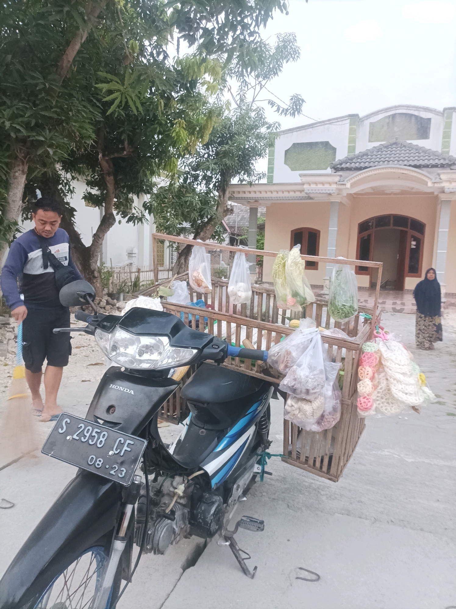In Canga'an, Indonesia, a man stands next to a motorcycle equipped with a wooden rack carrying various goods. He is wearing a long-sleeved shirt and shorts, and is holding a broom. The motorcycle has a license plate that reads "S 2958 CF 08-23". In the background, a house with a light-colored facade and a tiled roof is visible. A woman in a dark hijab and patterned skirt is standing near the entrance of the house. The scene appears to be outdoors, with trees and a clear sky overhead. The overall atmosphere suggests a daily scene of commerce or preparation for sales.