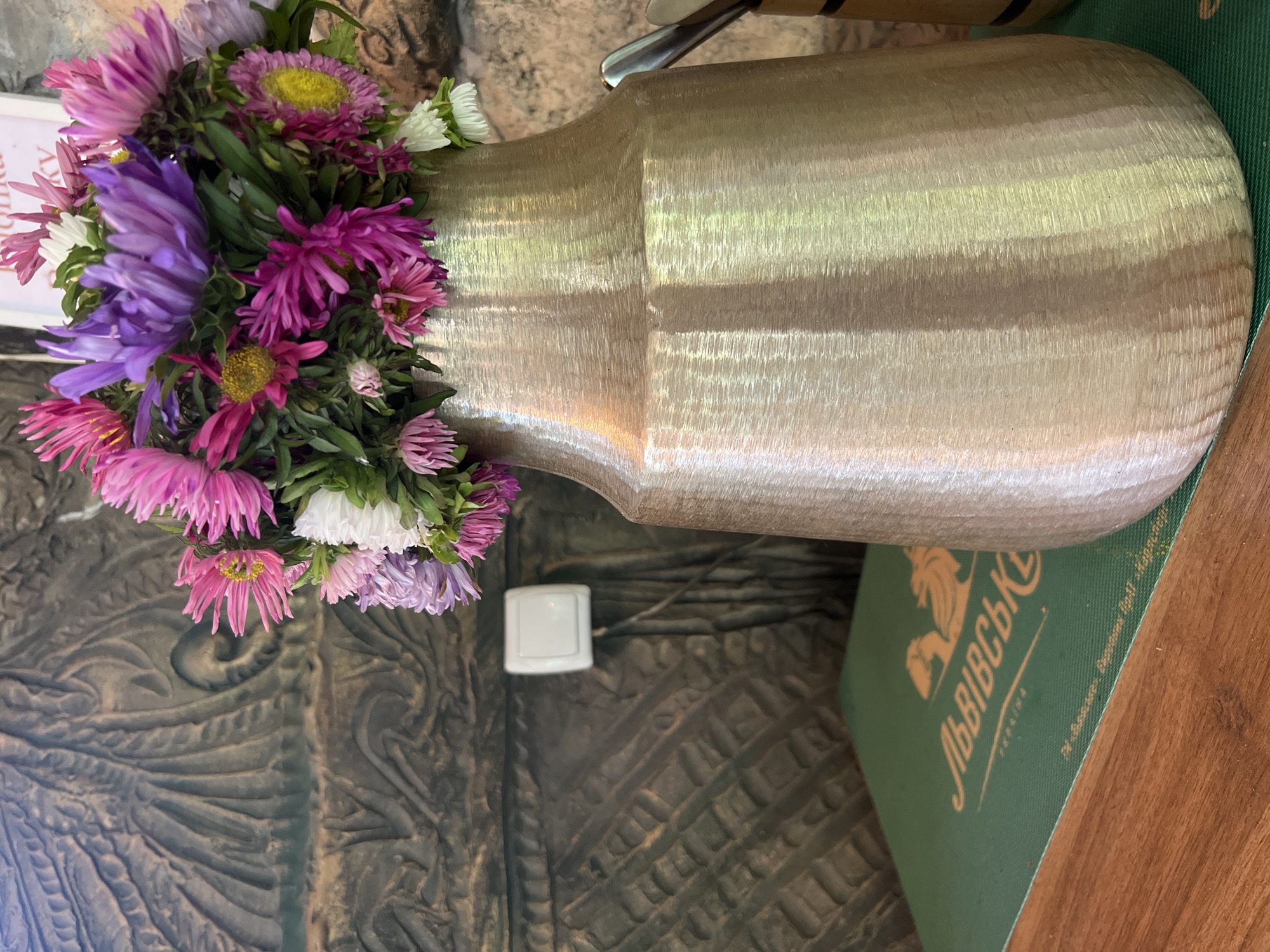 The image shows a close-up view of a bouquet of asters in a brushed metal vase, placed on a wooden table in what appears to be an indoor setting. The flowers are a mix of pink, purple, white, and yellow hues. Behind the vase, a textured wall with a decorative pattern is visible, along with a white electrical switch and outlet. In the foreground, a green placemat with orange text and a lion logo is partially visible, suggesting the location is a restaurant or cafe. The text on the placemat is in Ukrainian and reads "ЛЬВІВСЬКІ" which translates to "Lviv". Based on the context of this type of souvenir merchandise, the location is likely a cafe or shop in Yaremche, Ukraine.