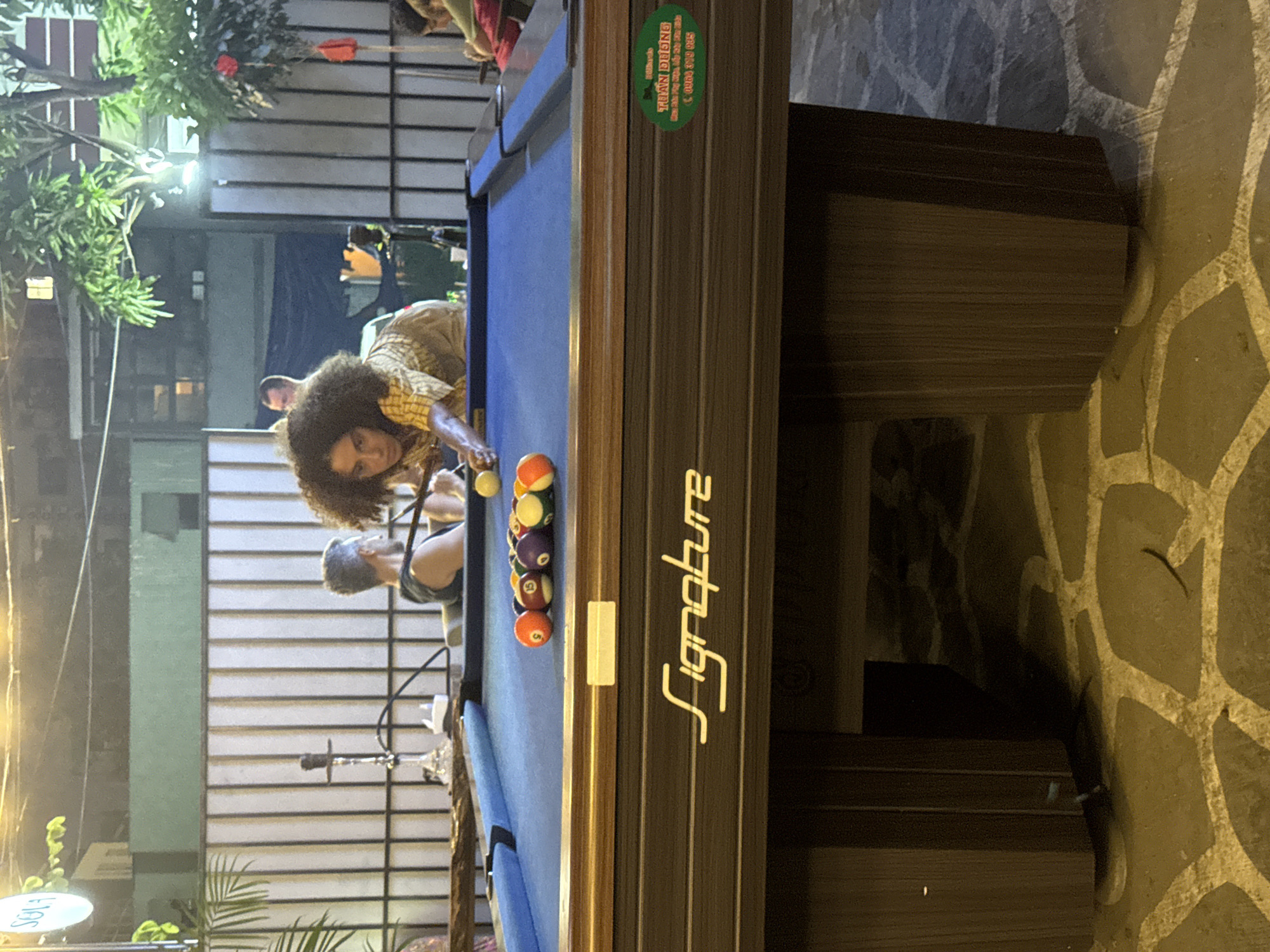 The image shows a group of people playing pool in an outdoor setting, likely in Đà Nẵng, Vietnam. A woman with an afro is leaning over a pool table, cue in hand, apparently aiming a shot at the racked balls. The pool table is branded "Signature." Another person, presumably her teammate or opponent, is standing next to her. The background includes a building with a slatted facade, greenery, and additional people standing or sitting nearby. The foreground features the pool table's wooden base and a stone-paved ground. A green sticker is visible on the corner of the pool table, potentially with Vietnamese writing and a phone number. The lighting suggests it is evening.