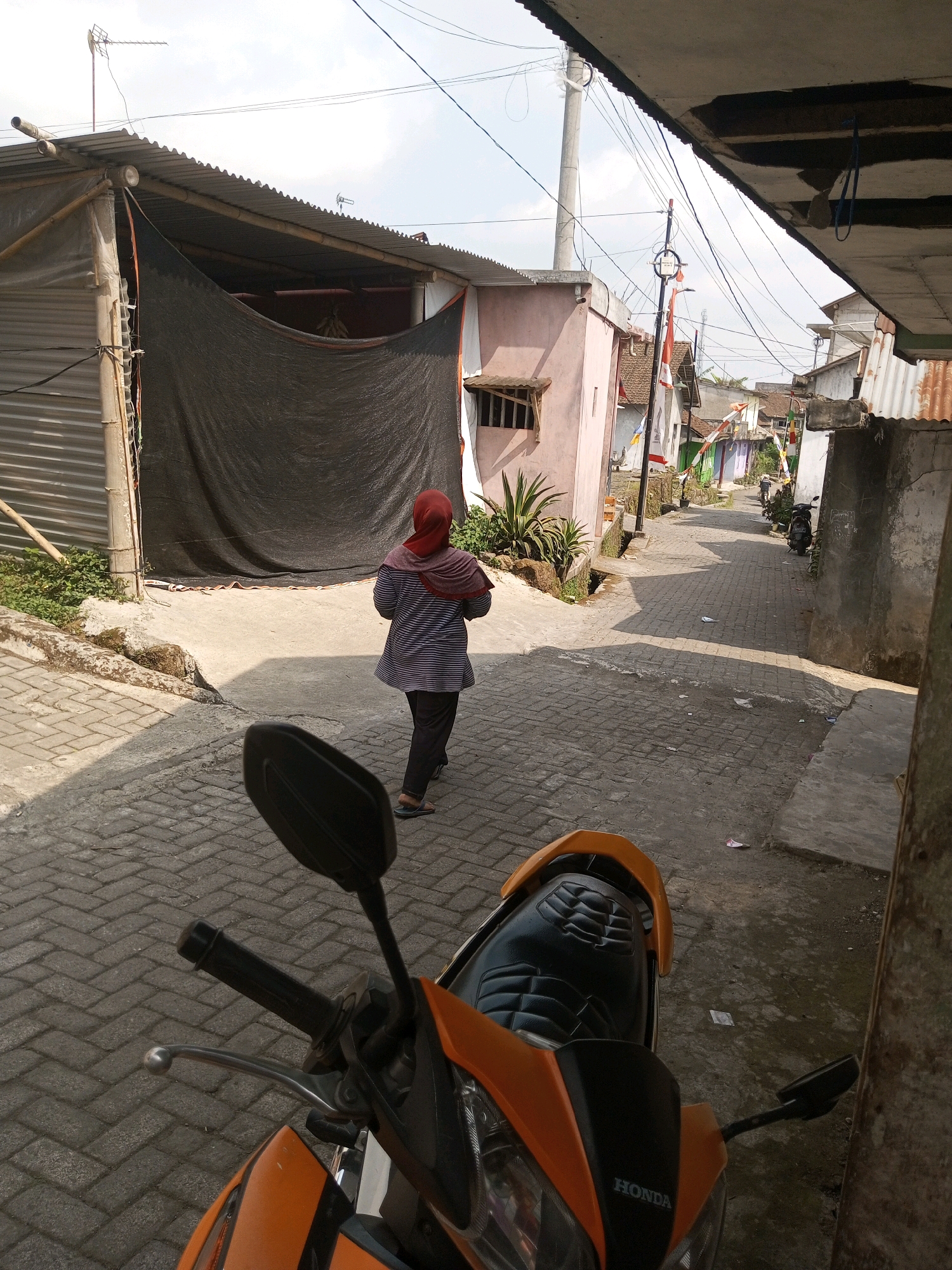 A person wearing a red headscarf and a striped shirt walks down a narrow, paved alleyway in Andongsili, Indonesia. The alley is lined with small buildings, some with corrugated metal roofs and others with pastel-colored walls. Indonesian flags are visible on some of the buildings, suggesting a local celebration or commemoration. A motorcycle is parked in the foreground, partially obscuring the view of the alley. The sky is overcast.