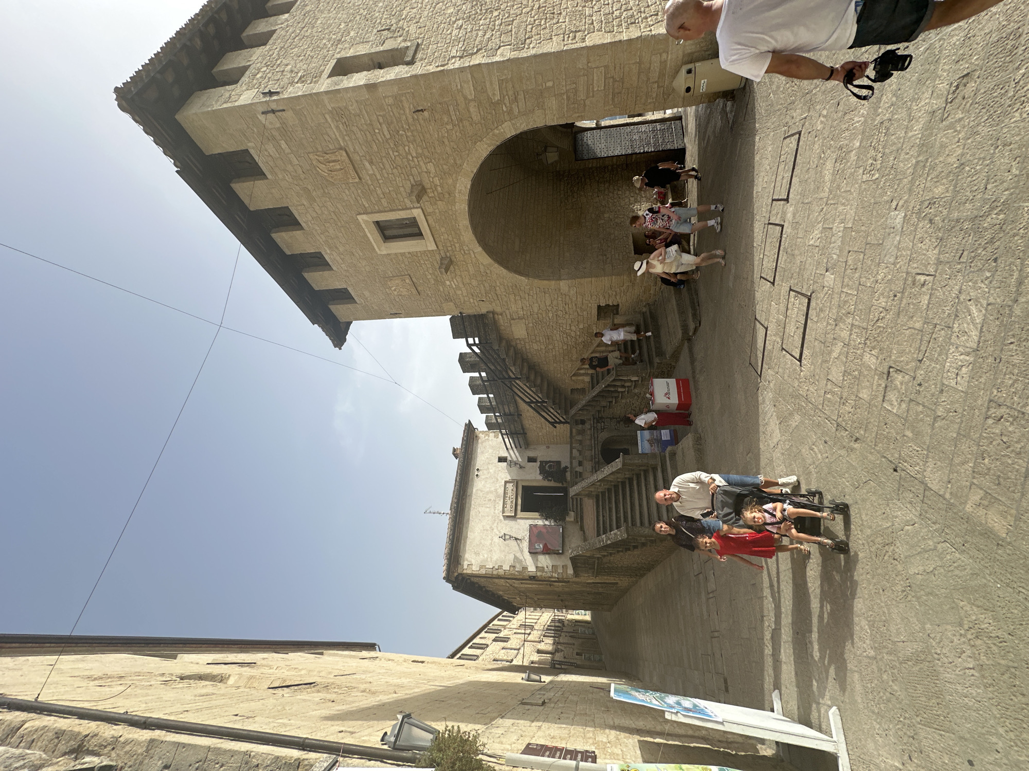 The image depicts a street scene in the historic center of the City of San Marino, San Marino. The perspective is from ground level, looking uphill and slightly to the right. The architecture is characterized by stone buildings with classic European design, some featuring crenellated battlements. A tall, arched gateway made of stone is prominent on the right side of the image, leading into a fortified structure.
In the foreground, a family is walking up the cobblestone street. A man is pushing a stroller containing a young child. A woman and an older child, possibly a girl in a red dress, are walking alongside them. Another man, partially visible from behind, is walking away from the camera to the right, holding a camera.
Further up the street, on the left, there is a building with a sign that reads "MUSEO TORTURA" (Torture Museum), with steps leading up to its entrance. Several other people are visible walking or standing in the background, some near the museum and others near the arched gateway. The sky is clear and blue, indicating a sunny day. The street is paved with irregular stone blocks, and shadows are cast by the buildings and the people. The overall scene conveys a sense of historical tourism in a European city.