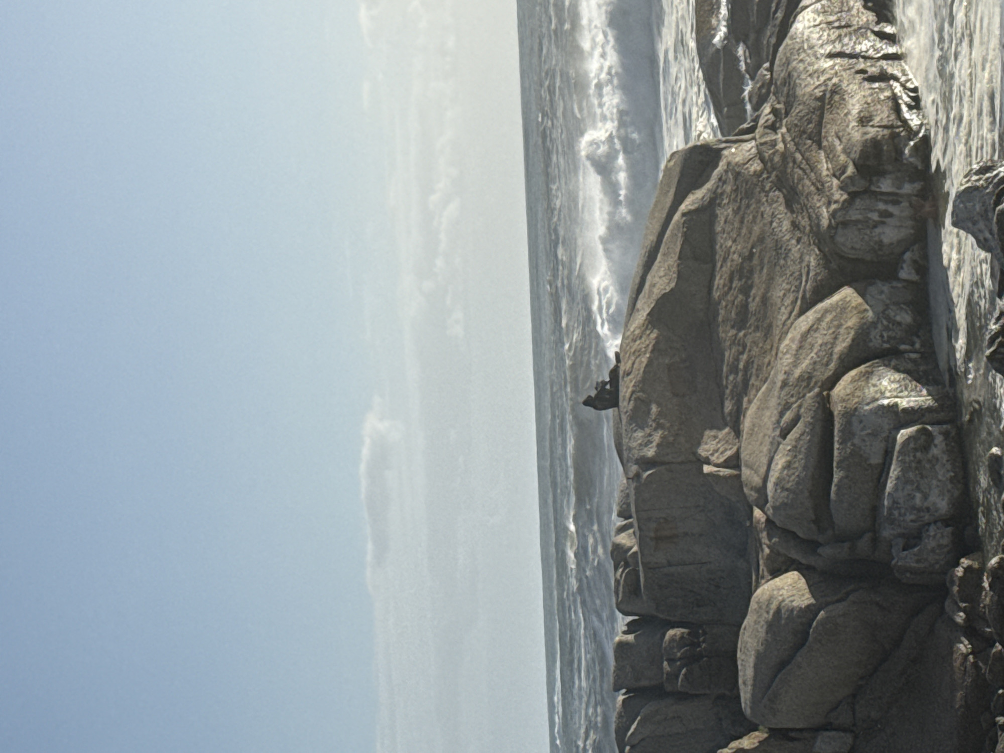 The image depicts a rocky coastline scene. To the right, a stack of grey rocks is visible, with the ocean appearing to crash against them. The water is churning and foamy, suggesting strong wave action. A solitary figure, silhouetted, stands on the rocks at approximately the center of the image. To the left, the sky is a pale blue with some light clouds. The horizon is visible, but obscured by the mist and the ocean's spray. While the exact location cannot be determined with certainty from the image, given the rocky coastline and light colored sky, it is plausible that the image was taken near the Cape Town, South Africa area.