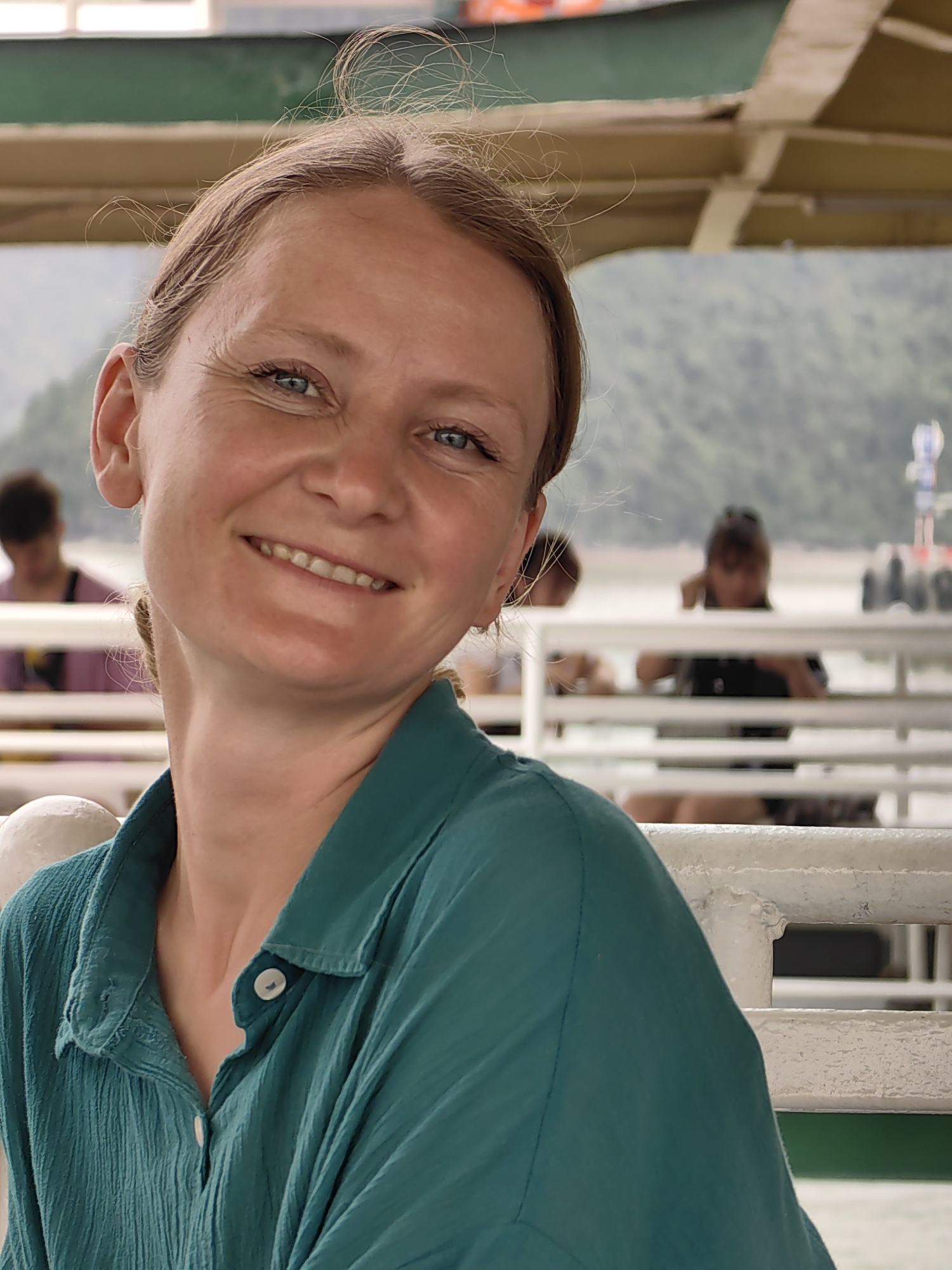 A woman with light skin and blue eyes smiles towards the camera. She is wearing a green button-down shirt. Behind her, other passengers are visible on what appears to be a boat or ferry. The background shows a body of water and tree-covered hills. A partial view of the roof structure of the boat is also visible. Given the location of Hải Phòng, Vietnam, the photo likely features Hạ Long Bay or a similar coastal location.