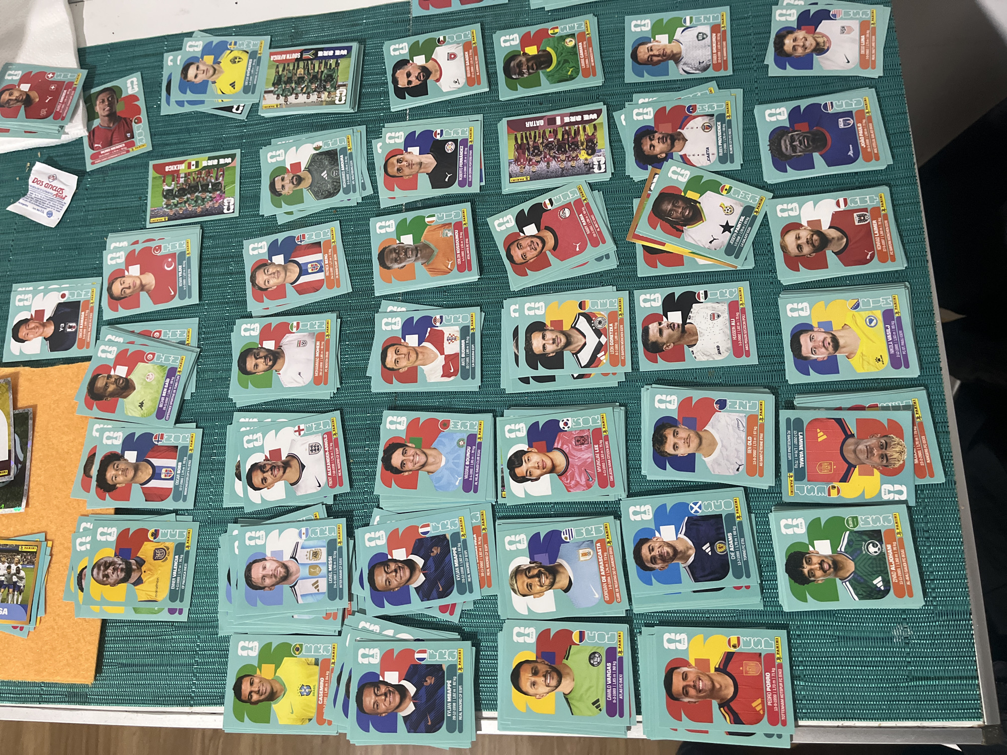 The image shows a collection of soccer player Panini stickers scattered across a teal-colored surface. There are numerous individual player stickers and a few team photo stickers. The stickers feature players from various national teams. Some stickers have been stacked together. A small white paper with text is visible in the upper left corner. The surface has a subtle grid pattern. The setting is likely indoors on a table or countertop.