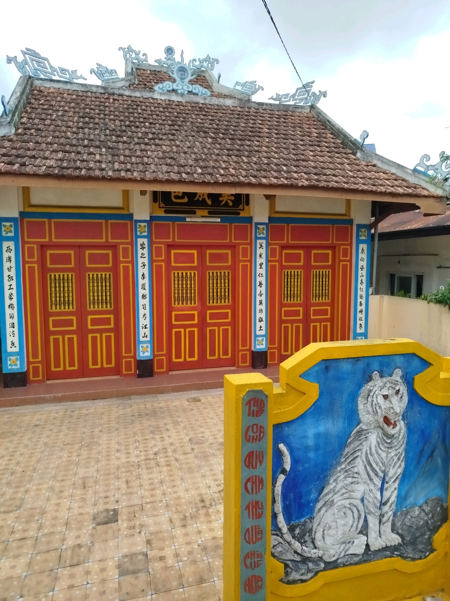 The image captures an outdoor scene of a building with traditional East Asian architectural elements, likely a temple or shrine, located in Da Lat, Vietnam. The building features a tiled roof adorned with decorative white ceramic figures. The facade is painted in vibrant colors, with large red doors accented by yellow frames and intricate grille work. Vertical blue panels with white inscriptions, likely calligraphy, flank the doors, and at the bottom of these panels are floral motifs.
In the foreground, a bright yellow structure with a blue background displays a relief sculpture of a white tiger sitting on a rock. This decorative element, which is partially in shadow, includes vertical red inscriptions on its left side. The ground in front of the building is paved with square tiles in a beige and grey checkered pattern. The sky above is overcast, with a visible power line crossing the frame. No people are visible in the image, and the overall impression is one of religious or cultural significance within a Vietnamese urban setting.