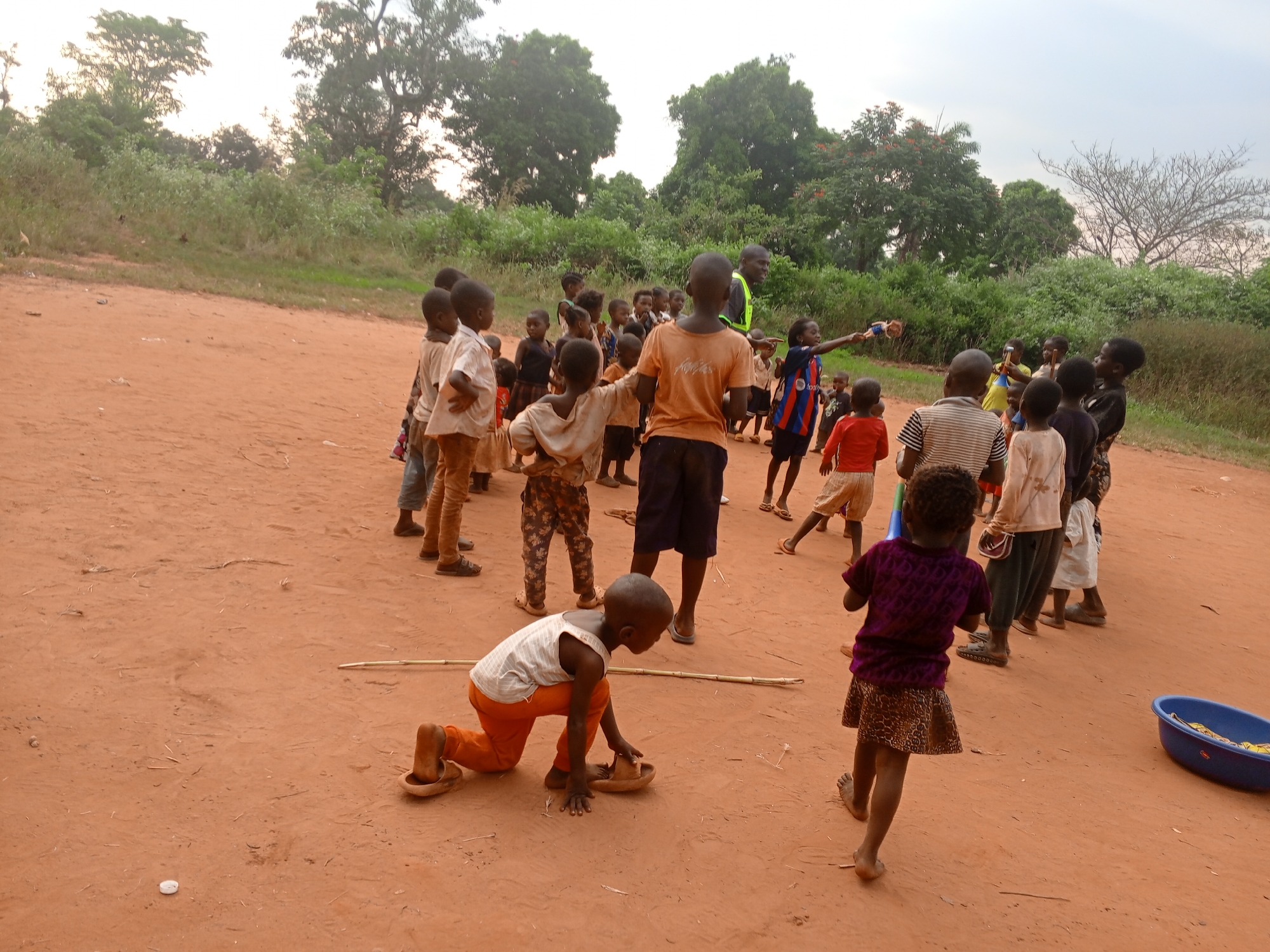 In Ngalula, Democratic Republic of the Congo, a group of approximately 30 children are gathered on a red dirt ground. Most children are standing and facing towards the center of the group, forming a semi-circle. They are of various heights and wearing a mix of casual clothing. One child kneels on the ground, seemingly playing with a stick and wooden sandals. A blue plastic tub sits on the right edge of the frame, containing yellow objects. Mature trees and dense vegetation line the background. The sky is overcast.