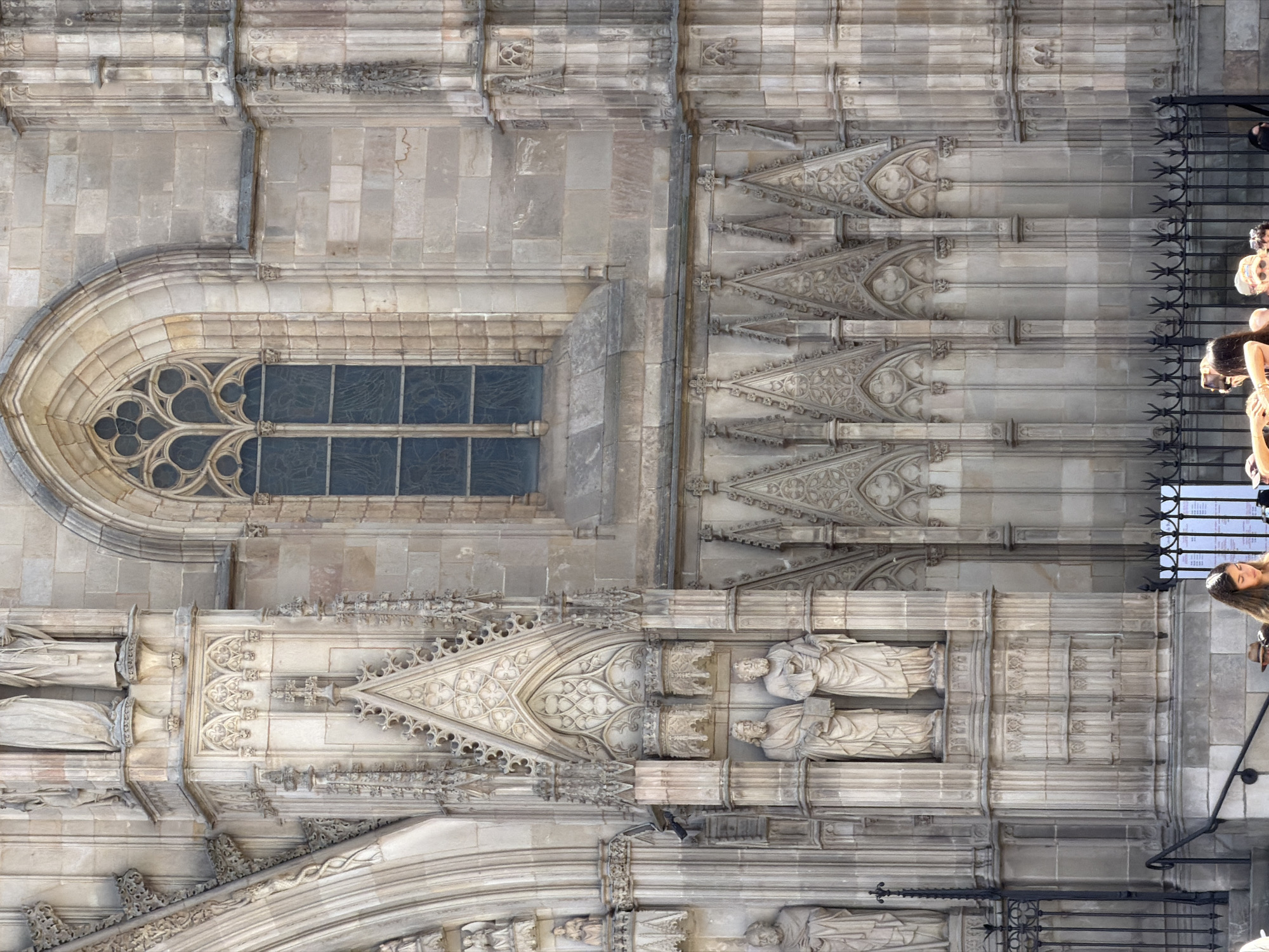 The image shows a close-up view of the facade of the Barcelona Cathedral in Barcelona, Spain. The cathedral features intricate Gothic architecture with detailed stone carvings and a large rose window. In the foreground, a group of people are gathered, appearing to be tourists taking photographs and looking at the building. The architecture includes statues of figures in niches, ornate pinnacles, and flying buttresses. The lighting suggests it is daytime, with shadows cast by the sun on the stone structures.