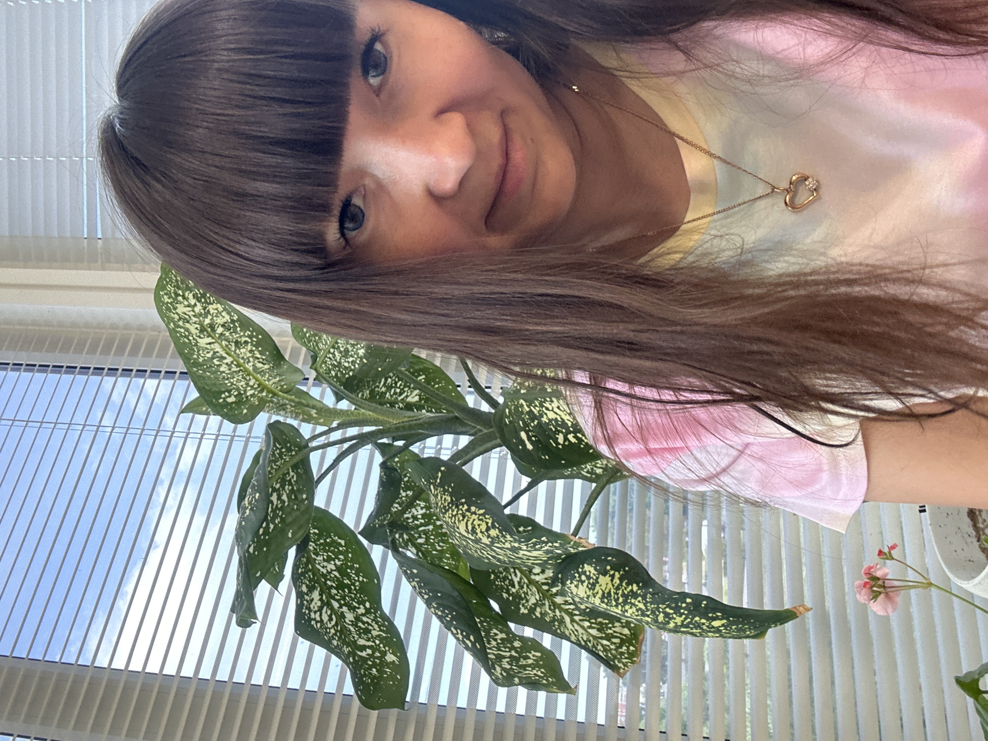 A woman with brown hair and bangs smiles, facing the camera at an angle. She wears a light-colored tie-dye shirt and a gold heart necklace. A plant with large green and white patterned leaves is in the foreground. Behind them, a window with blinds is visible, showing a view of a blue sky. The location is Chaikovsky, Russia.