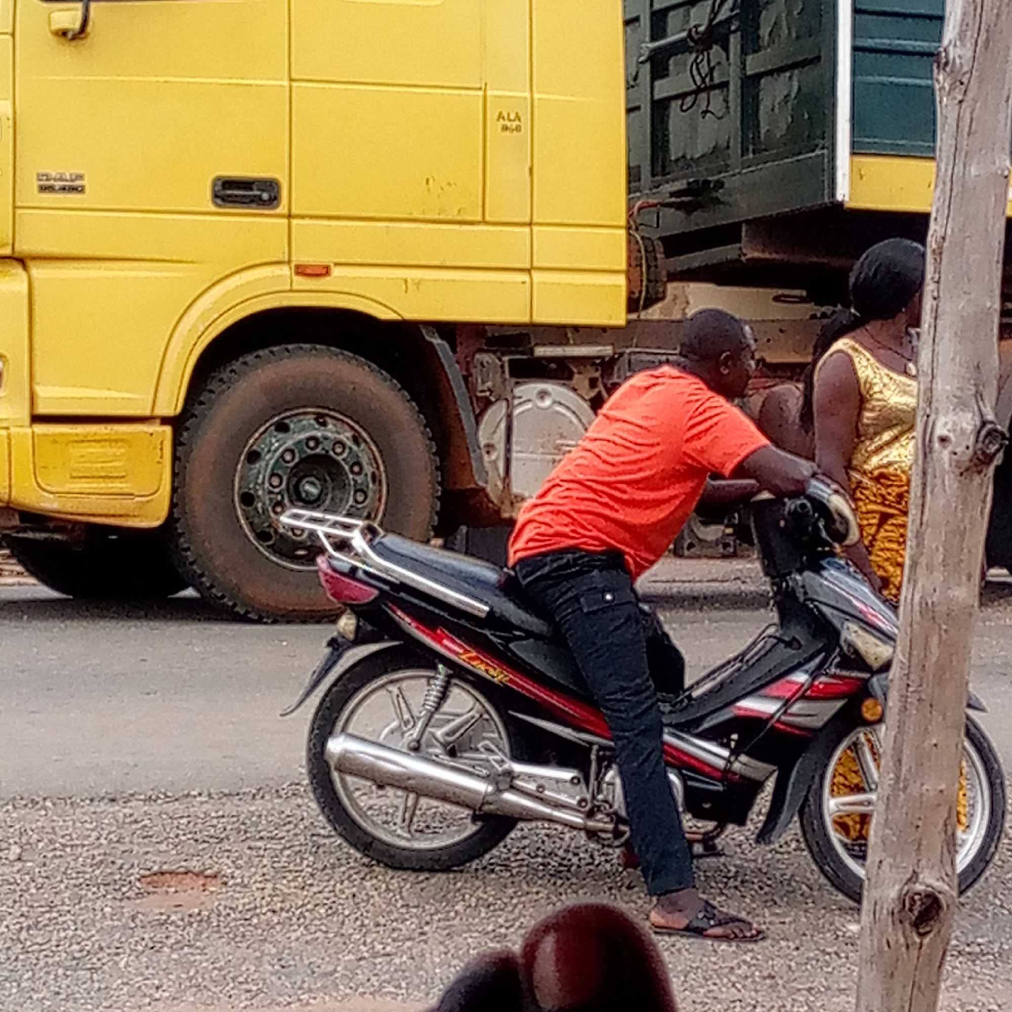 The image shows a man on a black motorcycle wearing an orange shirt and dark pants in Mararaba, Nigeria. He is leaning over the handlebars and appears to be interacting with a woman standing beside a large yellow truck. The truck has a dark green trailer or cargo area, and the ground is a rough, gravelly surface.