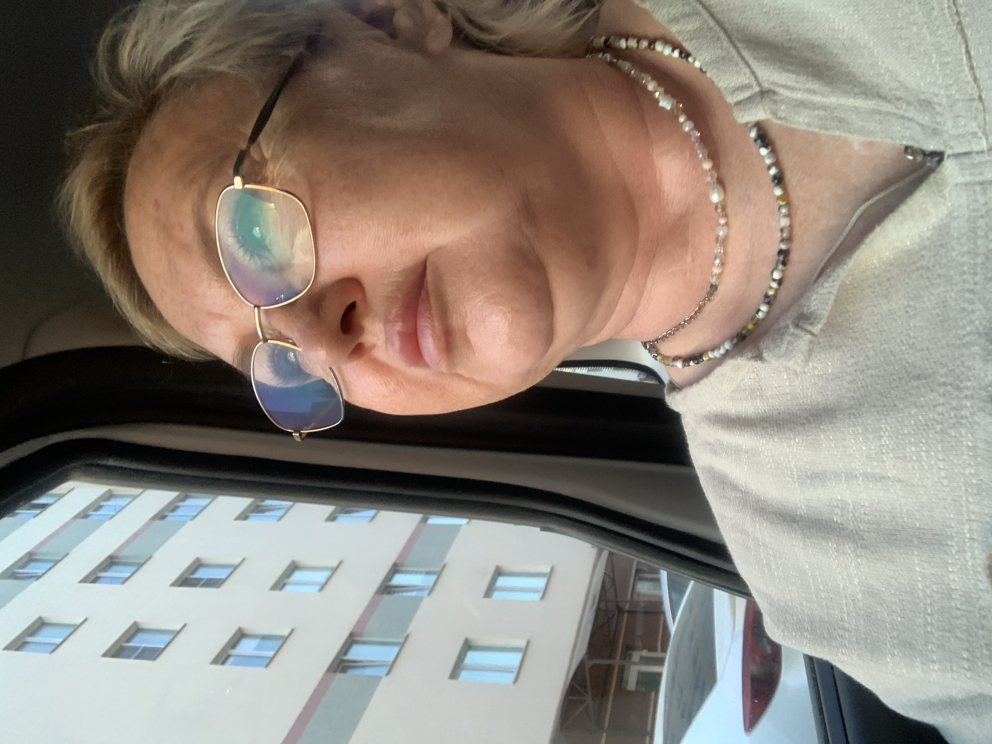 A photograph depicts a person with blonde hair and glasses, seemingly in a car, as evidenced by the window frame visible. The reflection in the car window indicates the person is in the city of Chaikovsky, Russia; reflected is a building with numerous windows. The person is wearing a light-colored garment and a multi-strand necklace. The person is looking downwards towards the car window. A partial view of a white vehicle's rear can be seen adjacent to the car.