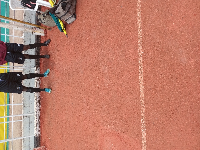 Two individuals in athletic attire stand on the edge of a red running track, with a white lane marker visible nearby. The individuals are wearing black shorts and colorful shoes. A white chair, a bag, and a partially visible barrier are situated close by. The surface appears to be a synthetic track used for sports.