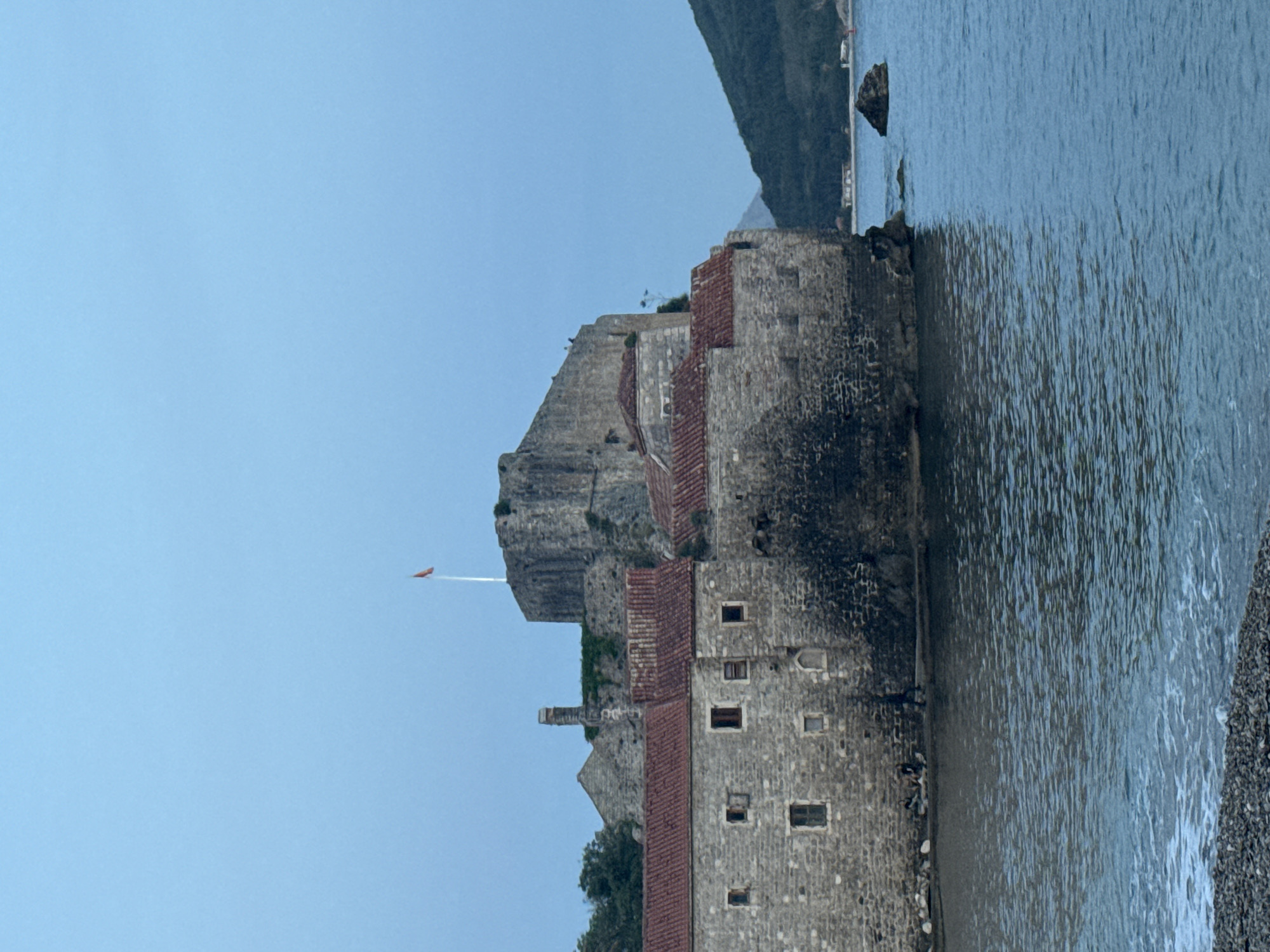 The image shows a stone fortress in Budva, Montenegro, with its walls extending into the sea. The structure has red-tiled roofs and several visible windows. A cliff rises behind the fortress. A flag is seen atop one of the towers. The Adriatic Sea laps against the fortress walls and a pebble beach is on the right side. A portion of the coastline is in the background, under a clear blue sky.