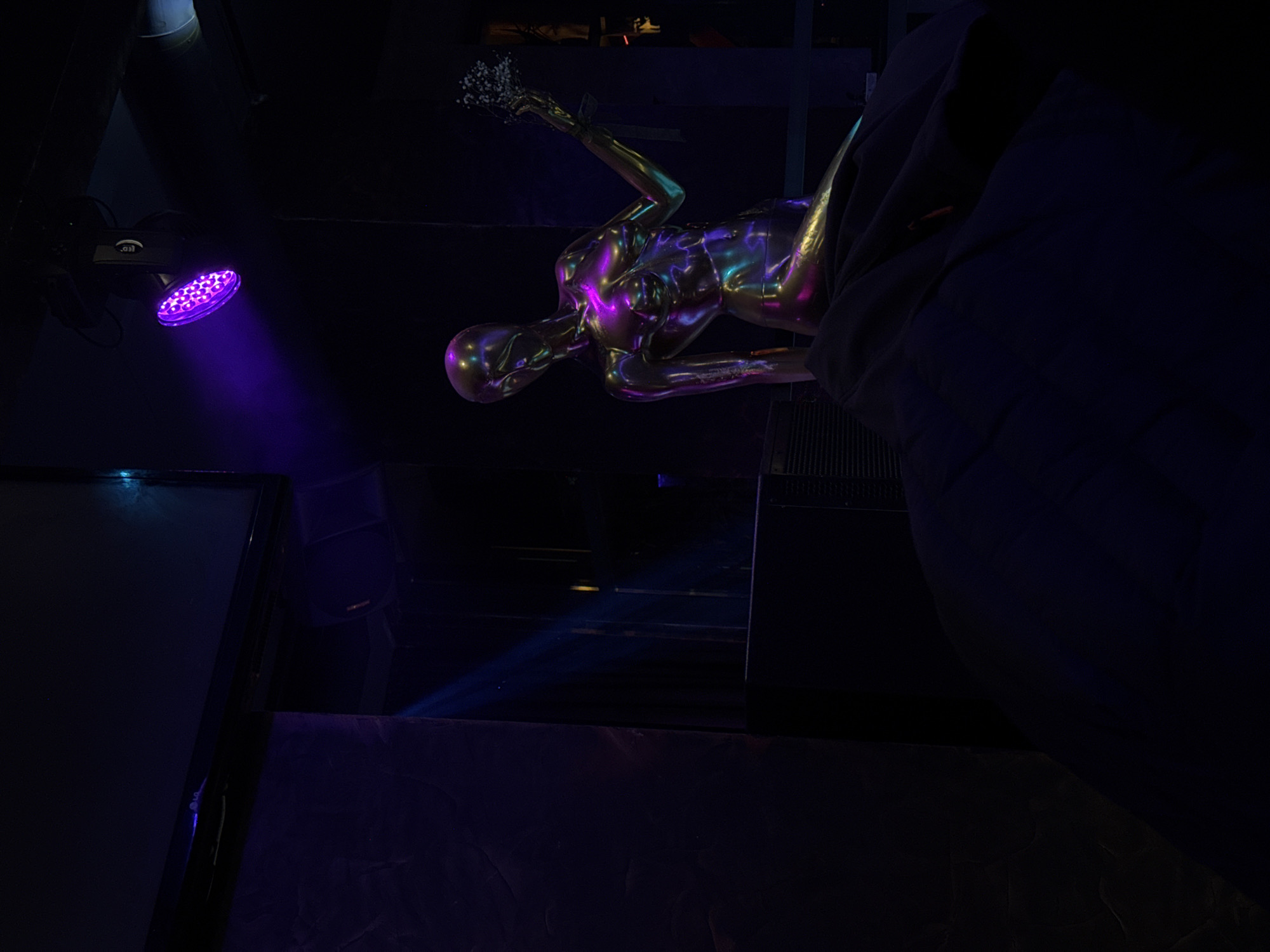 The image depicts a dimly lit interior, likely a nightclub or entertainment venue, in Burgas, Bulgaria. A metallic female mannequin lies reclined, illuminated with purple and gold lighting, holding a bouquet of flowers. A stage light casts a purple glow. Other objects such as a monitor or speaker appear in the scene. The environment is dark with only select parts of the image illuminated.