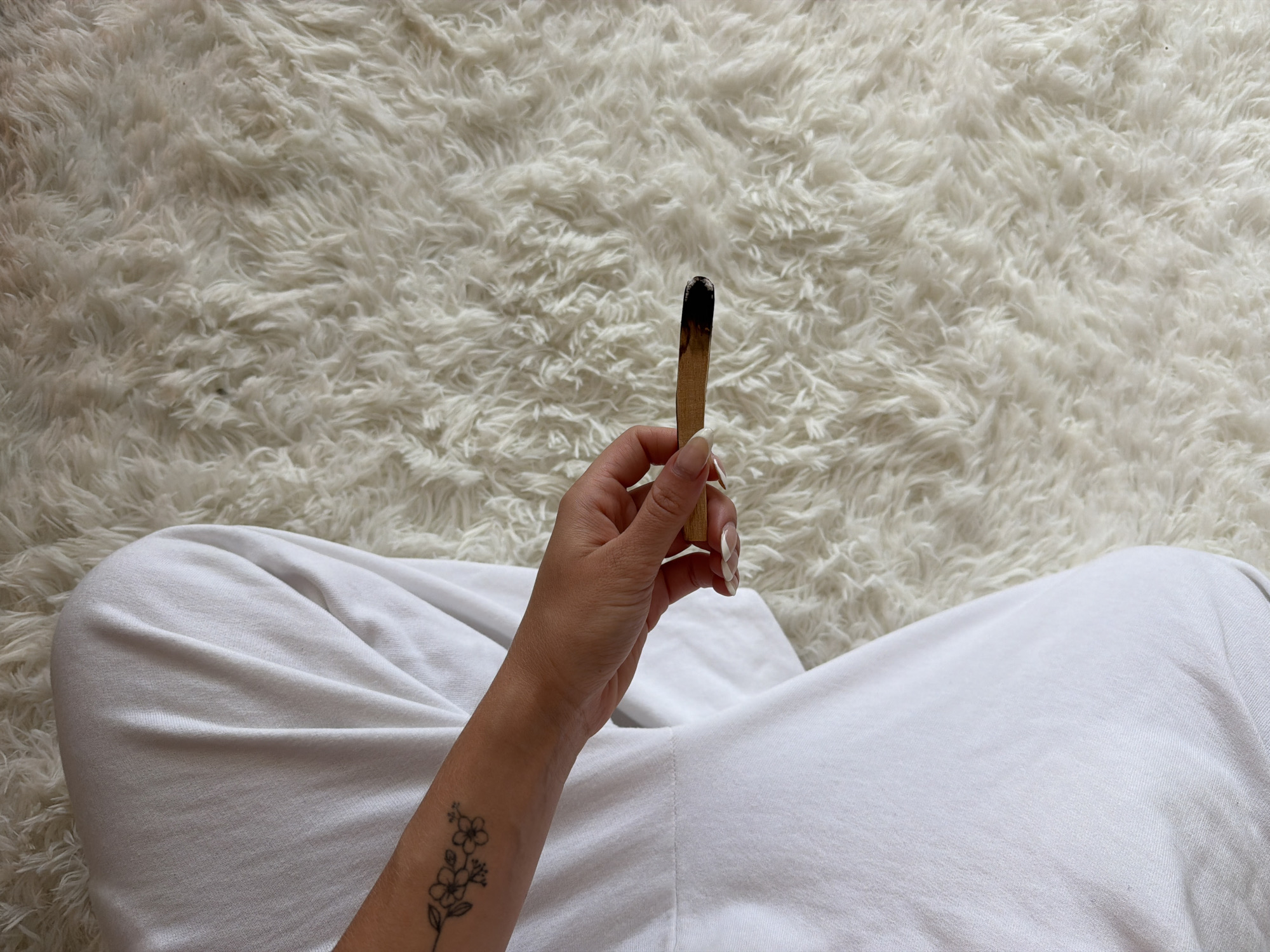 A first-person perspective image shows a person sitting on a white, textured surface, presumably a rug or bedding. The person is wearing white clothing, and their left arm is visible, displaying a small floral tattoo. They are holding a burnt Palo Santo stick in their hand. No definitive landmarks or details are present to confirm the location within Seoul, South Korea.