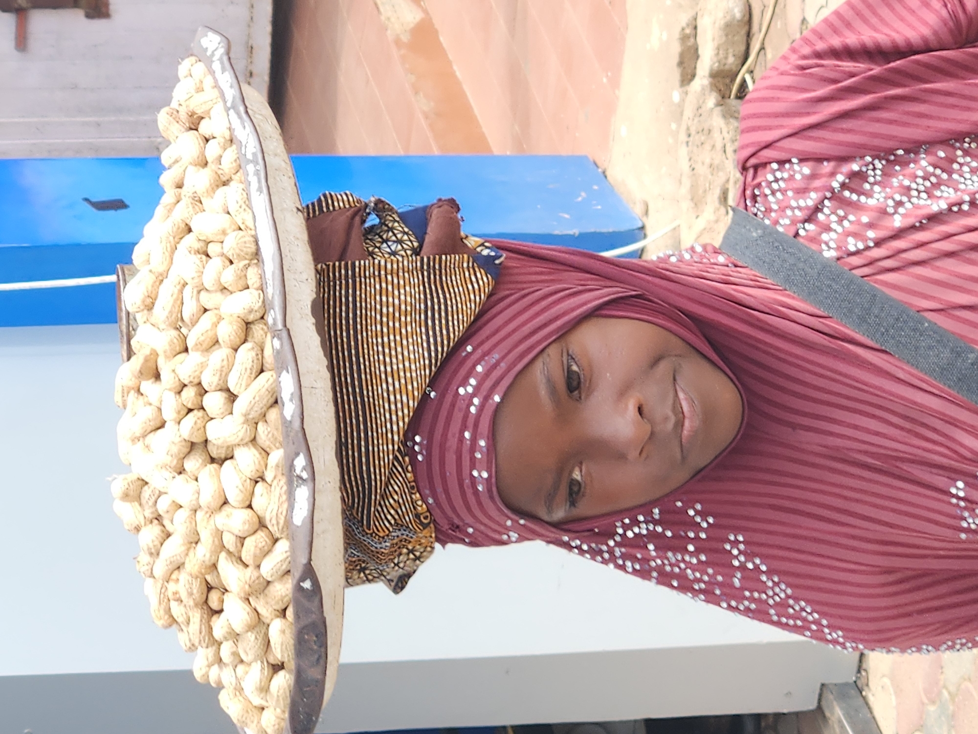 A young girl, wearing a maroon headscarf and a matching maroon tunic adorned with white beads, balances a large tray of peanuts on her head. The tray is a wide, shallow dish with a worn, dark rim, and it is filled with a mound of unshelled peanuts. Beneath the tray, several layers of fabric are visible, including a patterned cloth with horizontal stripes of yellow, brown, and black, and a dark red fabric. The girl's face is visible, with dark eyes and lips. A gray strap crosses her right shoulder. The background suggests an outdoor setting, with a blue structure on the right, a white wall on the left, and reddish-brown tiles or paving stones on the ground. The overall lighting indicates it is daytime.