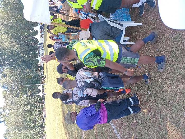 The image depicts an outdoor event on a grassy field, likely a sports ground, within Meru, Kenya. The precise location within the city is not identifiable from the visual information provided.
Approximately ten individuals are visible. In the foreground-left, a male wears a purple long-sleeved top, dark trousers with reflective strips, and a green cap, facing right. Centrally, a group of at least four males stands in a close formation; they wear matching patterned sports jerseys with numbers (e.g., "14" and "5" are visible) and dark shorts. One individual in this group wears orange athletic shoes.
To the right of this group, another male wears a neon yellow high-visibility vest over a green patterned short-sleeved shirt and dark patterned shorts. He raises his right hand and wears dark blue socks with dark grey slip-on shoes. Further right, under a white canopy tent, a male in a dark hoodie is seated on a white plastic chair. Other individuals are partially visible under or near this tent, including one in a yellow shirt and another in an orange top. Bags, possibly containing water, are visible near the seated individual.
The ground is a grassy field with some visible dry patches and a white field marking on the far left. In the mid-to-far background, multiple white canopy tents are erected across the field. Two tall vertical poles, consistent with goalposts, are visible in the background. A dense line of tall trees forms the distant horizon. The scene is illuminated by natural daylight.