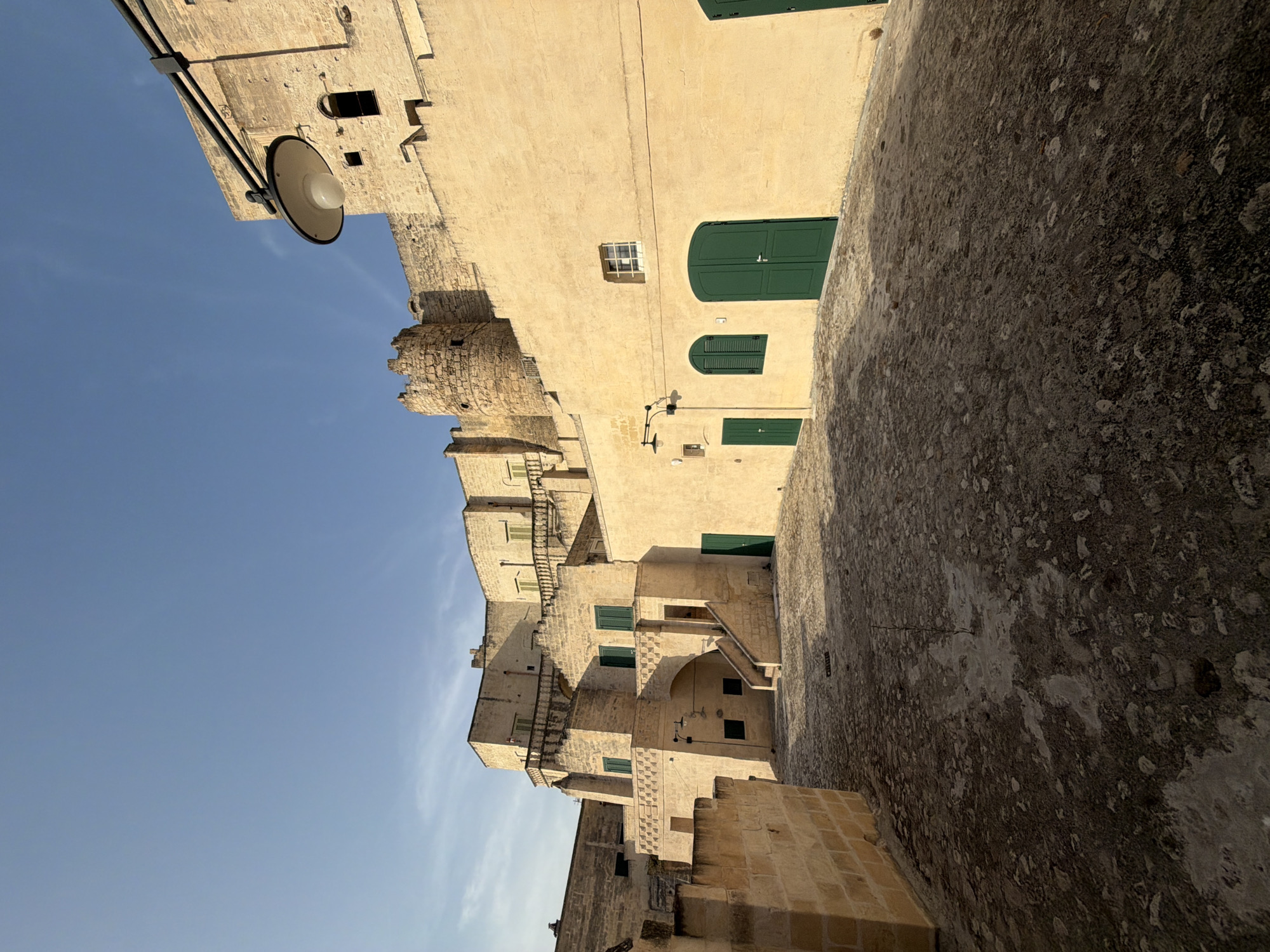 The image shows an alleyway in the Sassi di Matera, a historic district in Matera, Italy. The alley is paved with rough cobblestones and is lined with ancient stone buildings. The buildings are made of light-colored stone and feature numerous doorways and windows, many of which are covered by green shutters. Some buildings have multiple levels, with balconies and staircases visible. A large, circular stone tower, likely a defensive structure, is visible in the background, integrated into the buildings. A street lamp hangs from a metal pole extending from one of the buildings. The sky above is clear and blue, with a few wispy clouds. The sun is casting shadows, indicating it is likely late afternoon or early morning. There are no people visible in the image.
