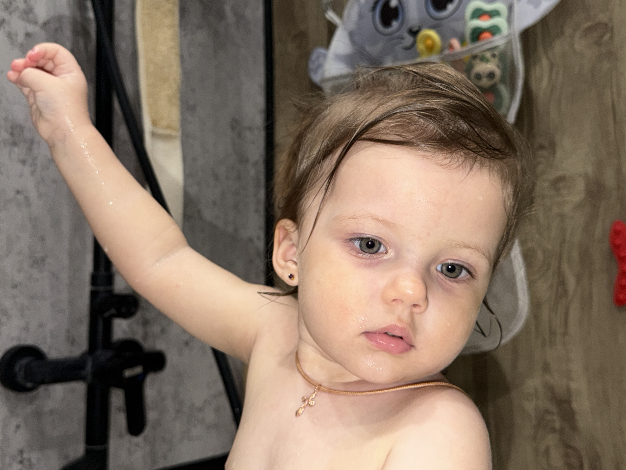 A young child, possibly a toddler, is depicted indoors, likely in a bathroom or shower area. The child, who appears to be fair-skinned with light brown hair and grey eyes, is wearing a small gold necklace with a cross pendant and small red studs in their ears. Their skin appears damp, suggesting they may have just finished bathing. The child's right arm is raised, with their hand near a black shower fixture mounted on a grey tiled wall. In the background, there is a wooden wall and a soft storage organizer containing a few stuffed animals and a pacifier. There is no information available to determine if the location is Reshetnikovo, Russia.