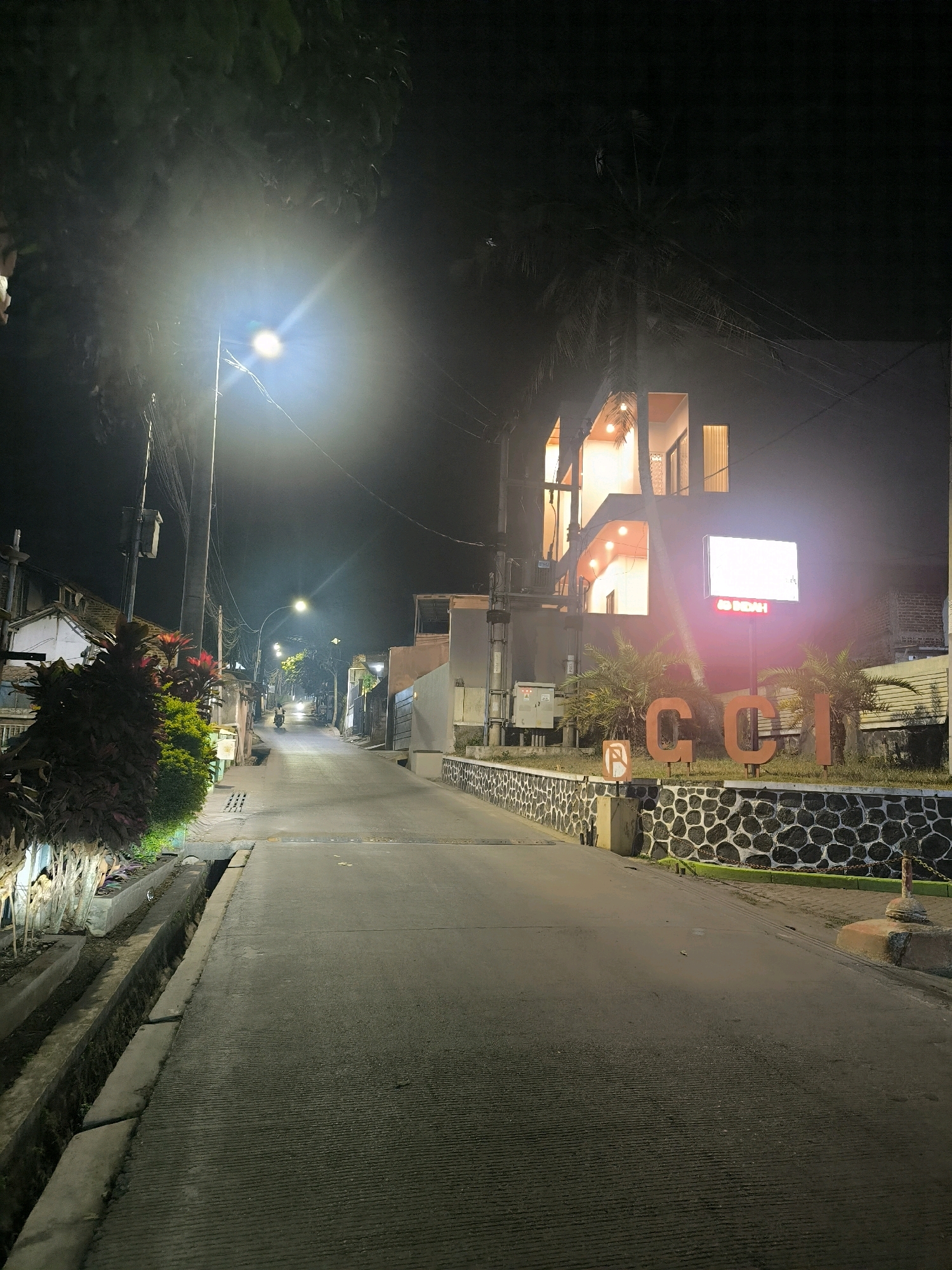 This is a night-time, eye-level, medium shot of a street in Bandung City, Indonesia. A lamppost illuminates the empty, paved road that slopes gently uphill. To the left of the road, a concrete drainage ditch runs alongside a row of plants, some of which are illuminated by artificial light. Further up the street, several buildings are visible, one of which features a modern architectural style with illuminated windows. To the right of the road, a stone wall with a decorative pattern runs along a property. In front of this wall, large orange letters spell out "CCI", and a rectangular illuminated sign is mounted on a pole above them. A single motorcyclist is visible in the distance, traveling up the street. The sky is dark, with the exception of the bright glare from the lamppost and the artificial lights from the buildings.