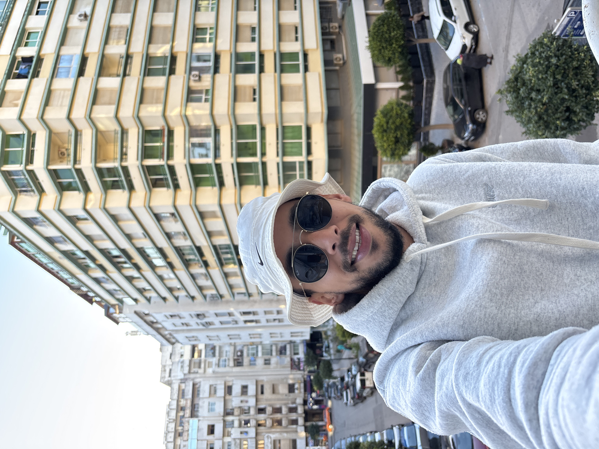 A male individual is captured in a selfie in Tanger, Morocco, smiling directly at the camera. He wears a white Nike bucket hat, dark round sunglasses with thin gold-toned frames, and a light gray textured hooded sweatshirt. The background features an urban environment. To the right, a multi-story building with a light yellow facade and teal-trimmed angular balconies is prominent, displaying numerous windows, some with external roller shutters, an air conditioning unit, and clothes hanging from an upper-floor window. To the left, a street curves, densely lined with parked cars, including an orange car visible further down the road. Behind the street, older, lighter-colored residential buildings rise. Small, spherical trees are planted along the right side of the street, where a lone pedestrian in dark clothing with a brightly colored head covering walks past parked vehicles. Green foliage is visible in the lower foreground. The sky is clear and light blue.