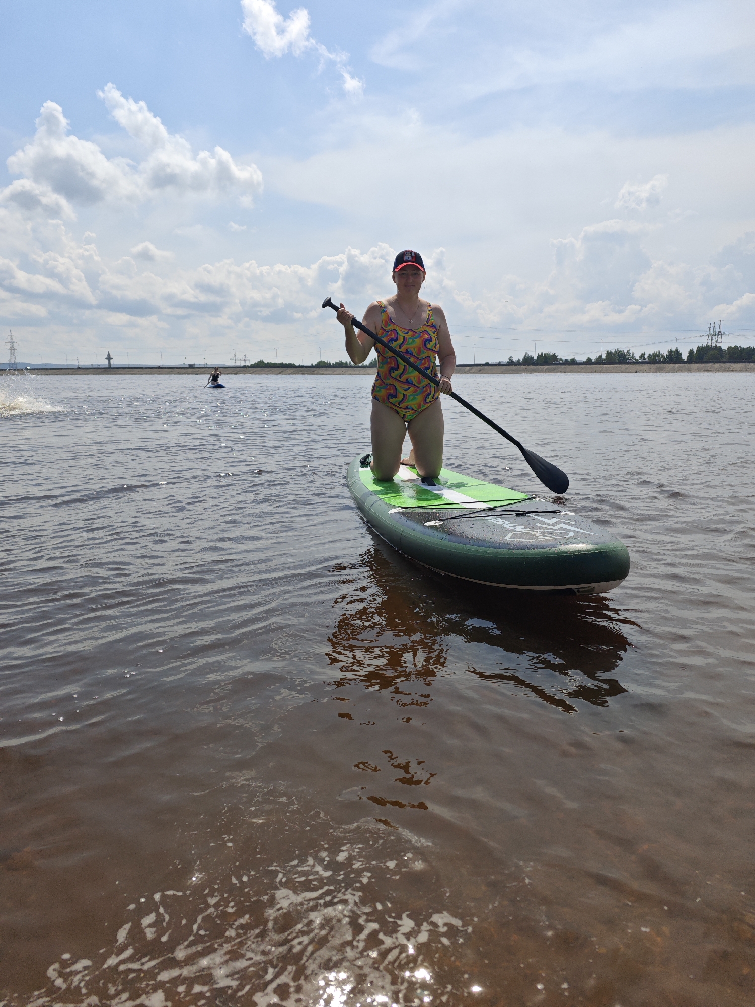 An adult woman in a colorful swimsuit and a baseball cap is kneeling on a green stand-up paddleboard in a body of water. She is holding a paddle with a black shaft and blade. Her left hand is on the paddle shaft, and her right hand is near the top of the shaft. She is looking towards the camera with a neutral expression. The paddleboard is green with white accents. The water is a reddish-brown color and appears calm with small ripples. In the background, there is a person on another paddleboard in the distance, and a shoreline with trees and power lines. The sky is blue with scattered white clouds. The scene is set in Noviy, Russia.