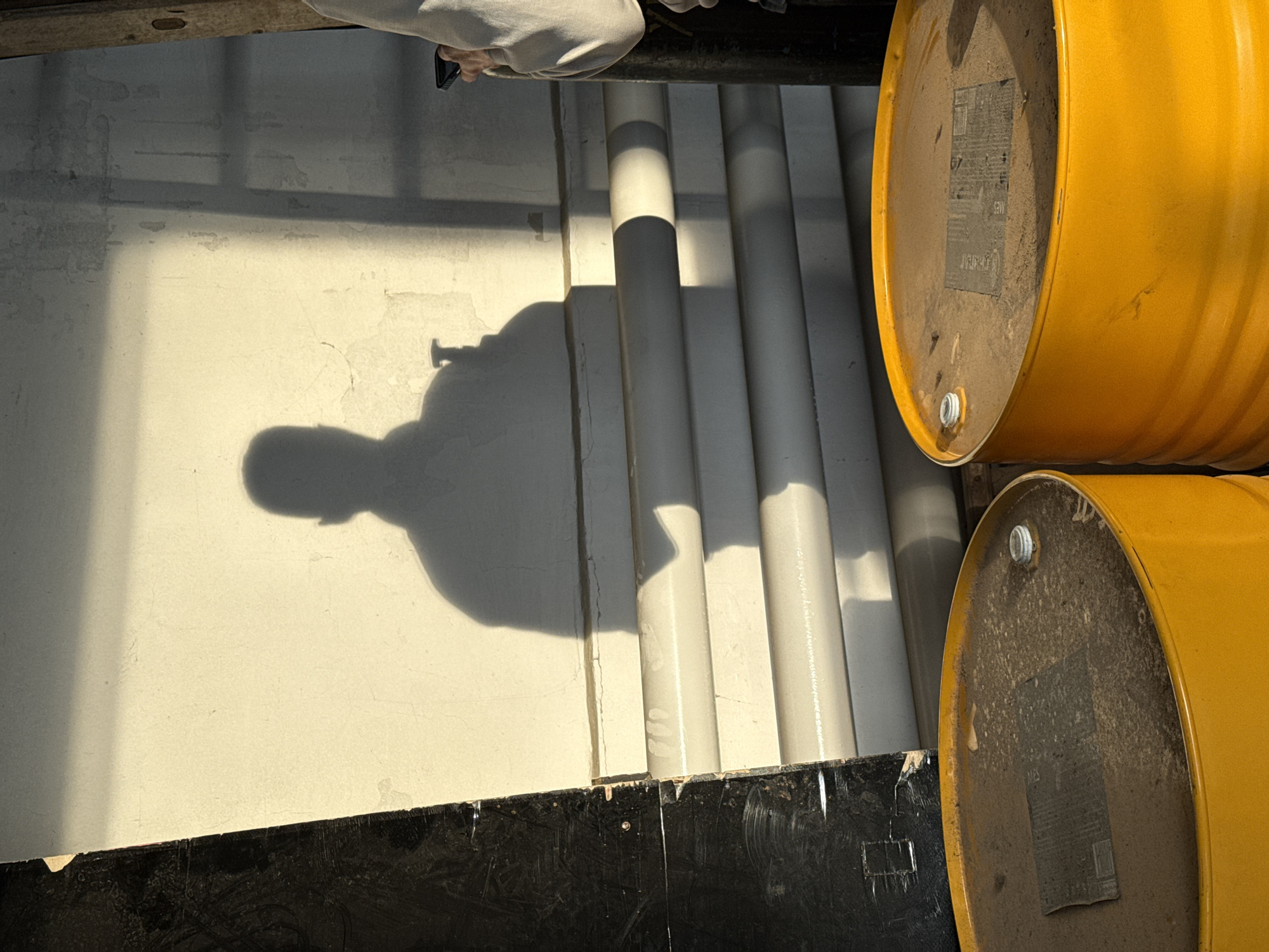 The image shows a shadow of a person holding a phone cast on a white wall in an industrial setting. Adjacent to the wall are four light gray cylindrical pipes. Two yellow barrels are positioned on the right side of the image, close to the pipes. The barrels have signs on the ends. The scene includes a black border along the bottom of the wall. The location is implied to be Chaikovsky, Russia.