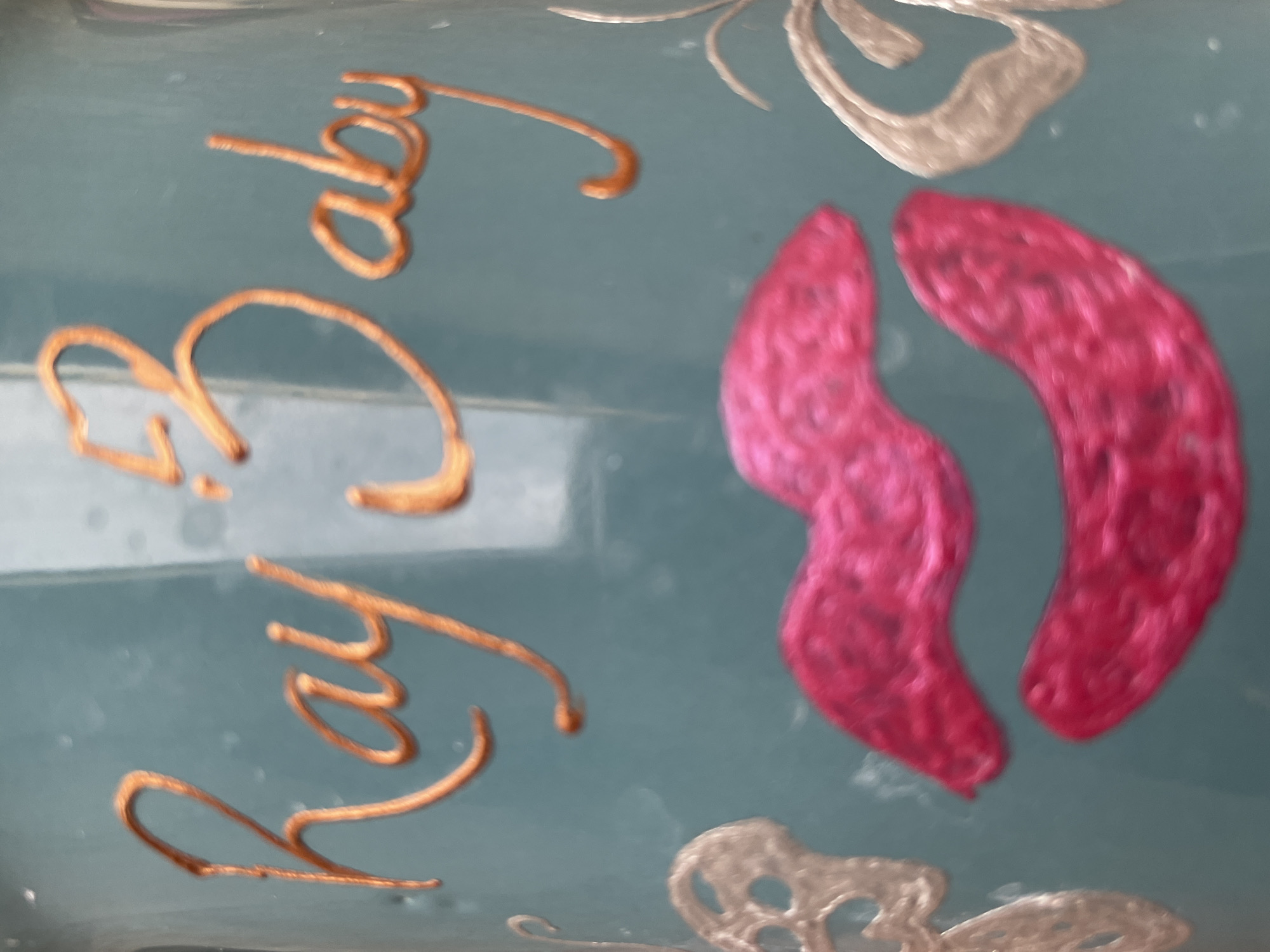 The image depicts a close-up of a pale blue surface decorated with raised embellishments. Two sets of cursive writing, likely the name "Ruby Baby," are rendered in a metallic gold color and rotated at an angle. Between these names are a pair of curvy, kidney-shaped designs filled in with pink texture, possibly representing a pattern of woven yarn or fabric. Two silver-colored butterfly shapes are partially visible at the top and bottom of the frame. The image appears to be taken indoors under diffuse lighting. Narayi, Nigeria, is not identifiable in the image.