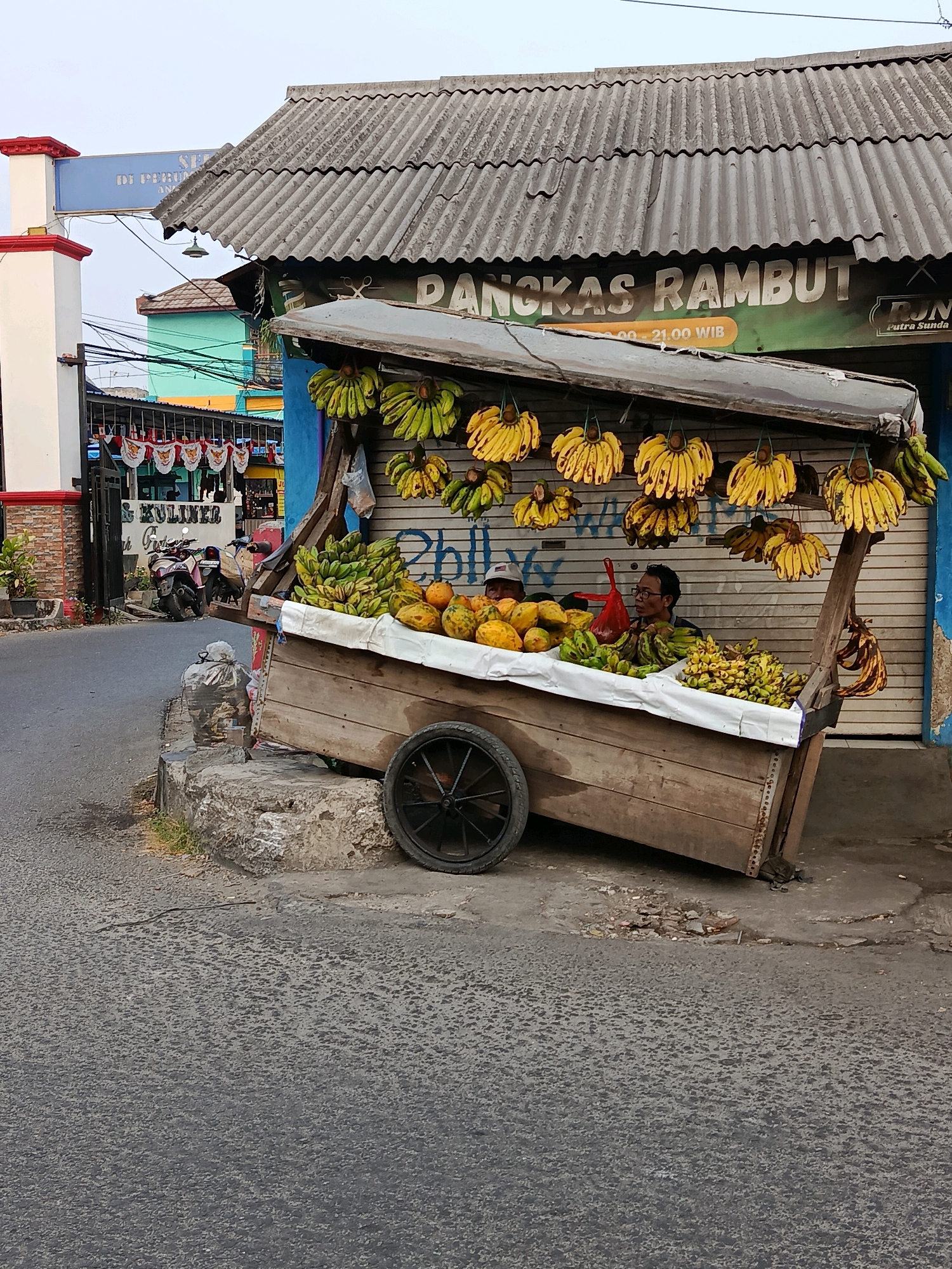 The image shows a fruit stall on a street in Bekasi, Indonesia. The stall is a wooden cart filled with various fruits, including bananas, papayas, and other tropical fruits. The cart is set up on the side of a road, with a corrugated metal roof providing shade. Two men are visible behind the fruit stall, likely the vendors. In the background, there are buildings, some colorful, and a sign that reads "PANGKAS RAMBUT" (haircut) and another sign with a business name that appears to be "PUTRA SUNDA". The overall scene depicts a typical street vendor setup in an urban environment.
