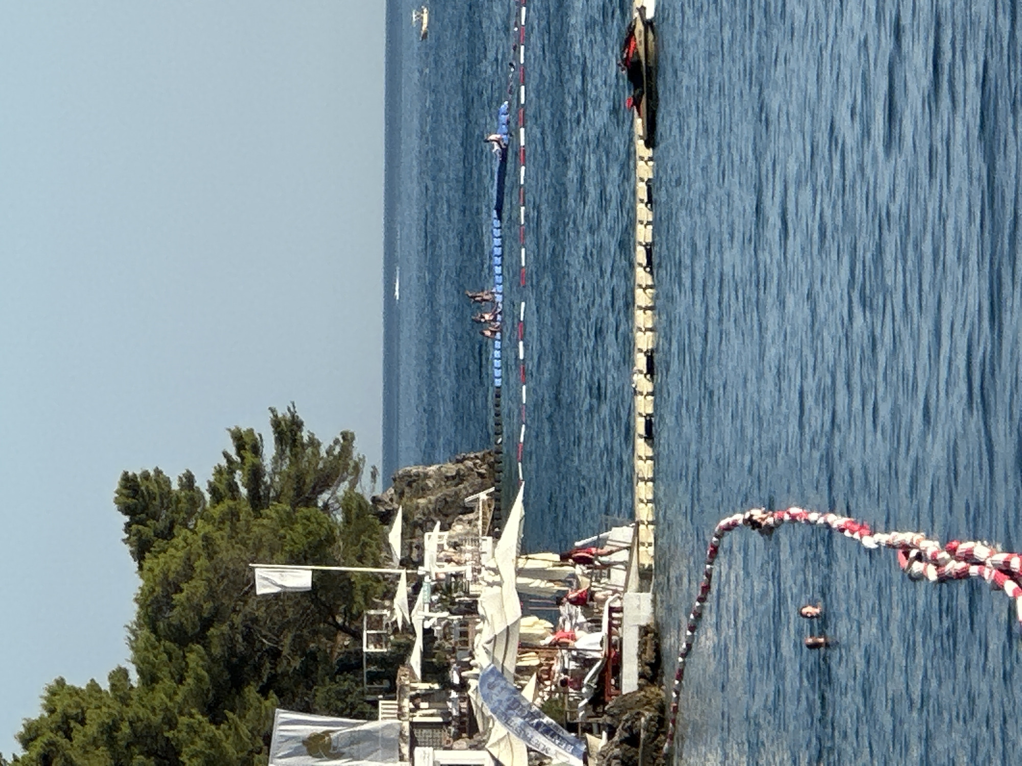 This is a photograph of a scenic waterfront area, likely a beach resort, in Budva, Montenegro, under a clear blue sky. In the foreground, the azure sea is marked by a rope barrier with alternating red and white buoys, delineating a swimming area. Two individuals are visible in the water, their heads above the surface.
Further out, a white floating platform structure stretches horizontally across the water. Along this platform, several people are engaged in various activities. Some are seated, while others appear to be standing. In the background, a blue floating walkway system is in place, with a group of people gathered on it. One individual is seen sitting on the edge of the blue walkway.
To the left of the image, a rocky shoreline is visible, dotted with lush green pine trees. Several white structures, possibly part of a restaurant or beach club, are built into the hillside, featuring white umbrellas providing shade. A white flag flutters from a flagpole. A jet ski is docked on a yellow floating platform near the right side of the frame. In the distance, a small white boat is visible on the horizon. The overall atmosphere is one of leisure and recreation in a Mediterranean coastal setting.