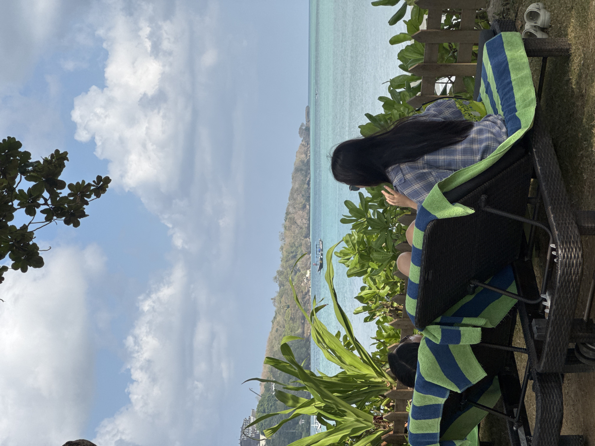 Two people are relaxing on lounge chairs on a grassy area facing the ocean. The lounge chairs have blue and green striped towels. The person in the foreground, on the right, is a woman with long dark hair wearing a blue and white plaid shirt. She is looking out at the ocean. The person on the left is partially visible, wearing dark clothing. In the background, the turquoise ocean stretches out to the horizon, with a hilly coastline visible in the distance. A small boat with an umbrella is on the water. Lush green foliage is in the foreground and along a wooden fence. The sky is blue with scattered white clouds. The scene appears to be a resort area in Kuta Selatan, Indonesia, likely a beach resort given the ocean view and lounge chairs.