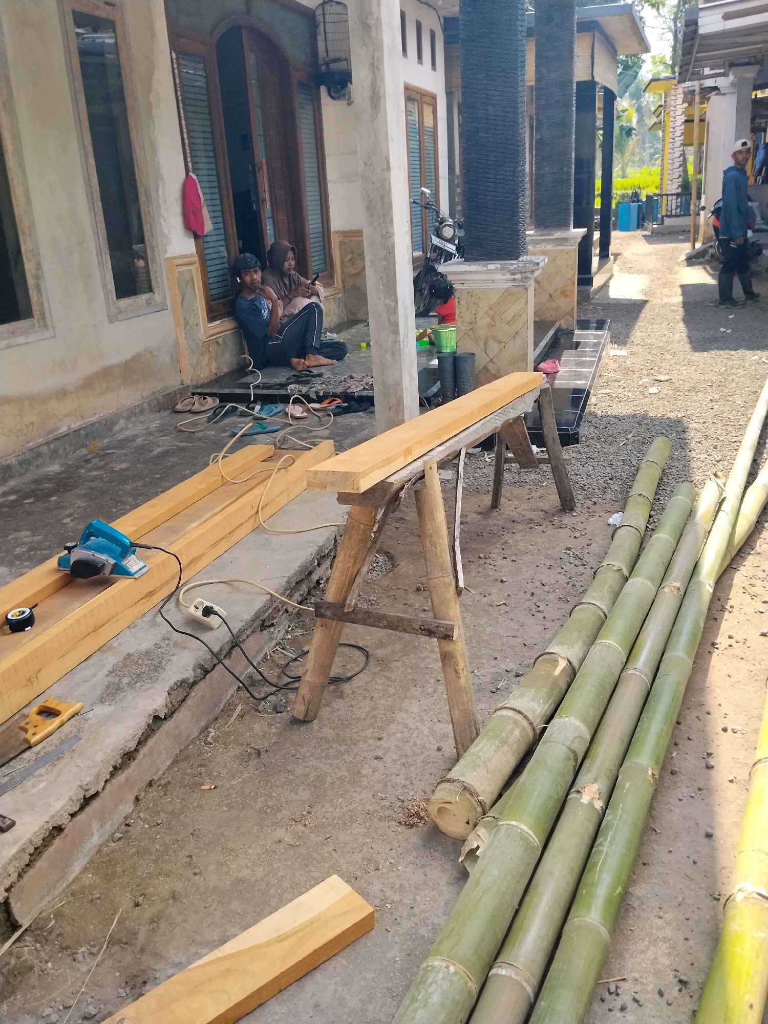 The image depicts a scene in Panggungan, Indonesia, likely a residential area undergoing some form of construction or woodworking. In the foreground, several large bamboo poles are laid out on the ground, with some lighter colored wooden planks also present. A woodworking tool, possibly an electric planer, is situated on one of the wooden planks, connected to an electrical outlet via a cord. Nearby, a pair of sawhorses supports another wooden plank.
In the background, two individuals, appearing to be young adults, are seated on the ground in front of a house. They are casually dressed, with one person wearing a headscarf. One of them is looking at a smartphone. The house has a prominent wooden door with glass panels and a birdcage hanging outside. The surrounding environment appears to be a mix of concrete and gravel ground, with some vegetation visible further in the background. A motorcycle is parked near the house. Another person, wearing a cap and a blue jacket, is standing further down the pathway, looking towards the right side of the frame. The overall impression is a candid shot of everyday life and work in a semi-rural Indonesian setting.