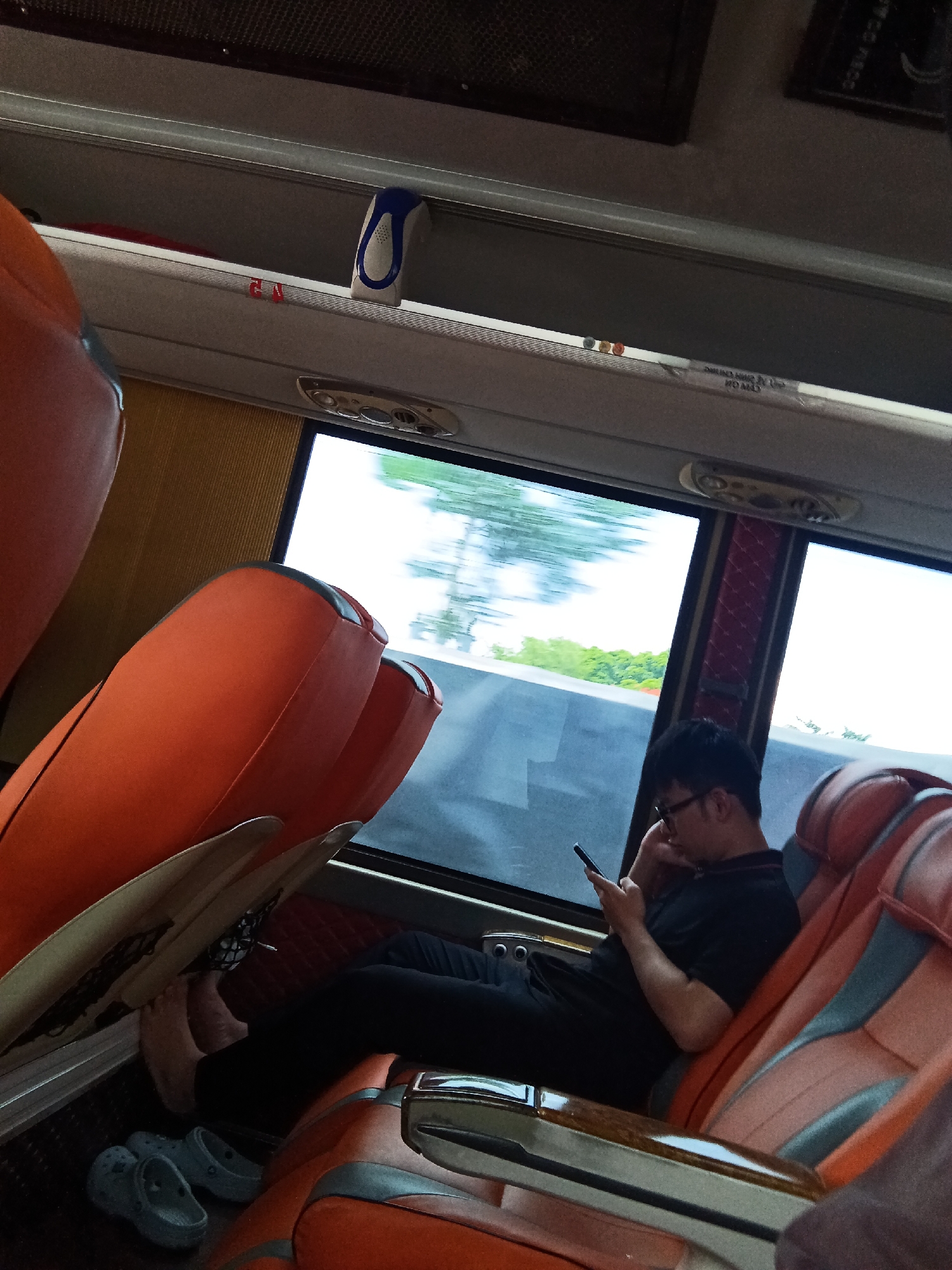 An overhead shot captures a man sitting on a bus, looking at his phone. He is wearing a black t-shirt and black pants, with glasses. He is seated in an orange and gray bus seat, with his feet resting on the floor. A pair of gray Crocs are also visible on the floor next to his feet. Through the bus window, trees and sky are visible, suggesting the bus is in motion. The bus interior is visible, with orange seats and a gray overhead compartment. There is a blue and white object attached to the overhead compartment, and red and yellow lights. The scene is dimly lit.