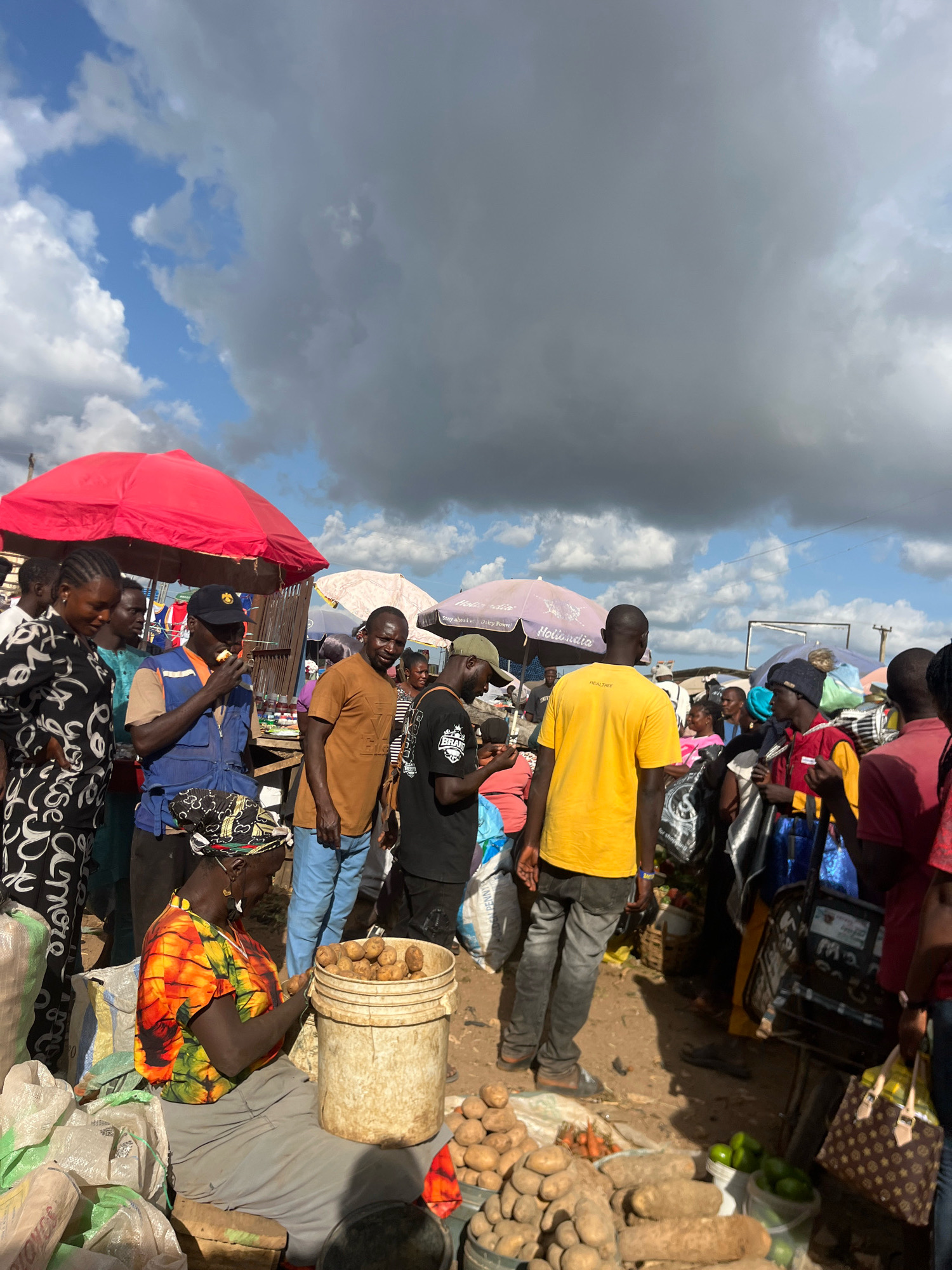 The image depicts a bustling outdoor market scene in Gura Top, Nigeria. A diverse group of people are engaged in various activities, suggesting a lively commercial environment. A woman in a vibrant, multi-colored shirt is seated, surrounded by sacks of produce and a bucket filled with potatoes, actively tending to her goods. Several individuals are standing and interacting, with some appearing to be customers and others vendors. The market is characterized by a collection of stalls with umbrellas providing shade, including a prominent red umbrella. Various items are displayed for sale, such as root vegetables like potatoes and what appear to be yams, as well as other produce and bags of commodities. The ground is dusty and uneven, typical of an open-air market. The sky above is partly cloudy, with a mix of dark and light clouds indicating variable weather conditions. The overall impression is one of a dynamic and active marketplace where commerce and community converge.