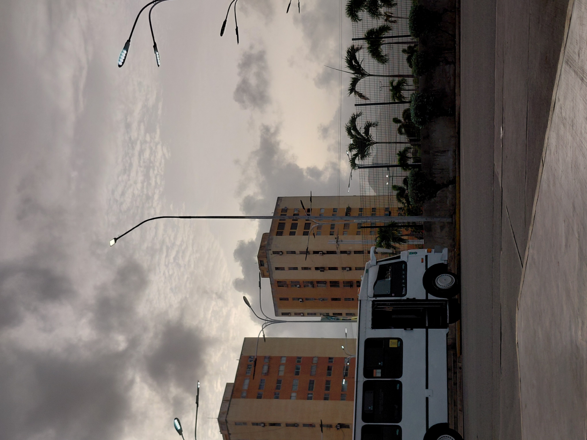 The image captures a street scene in Juan Griego, Venezuela, under a cloudy sky. In the foreground, a white bus with its doors open is parked on a paved road. To the right of the bus, a concrete barrier lines the edge of the road, behind which are several palm trees and dense green bushes. A chain-link fence stretches across the mid-ground, separating the road area from the palm trees and bushes. In the background, two tall, multi-story buildings with orange and yellow facades stand prominently. The buildings have numerous windows and appear to be apartment complexes or offices. Streetlights are visible throughout the scene, with some illuminated against the overcast sky. The overall atmosphere is dim and muted, characteristic of an overcast day.