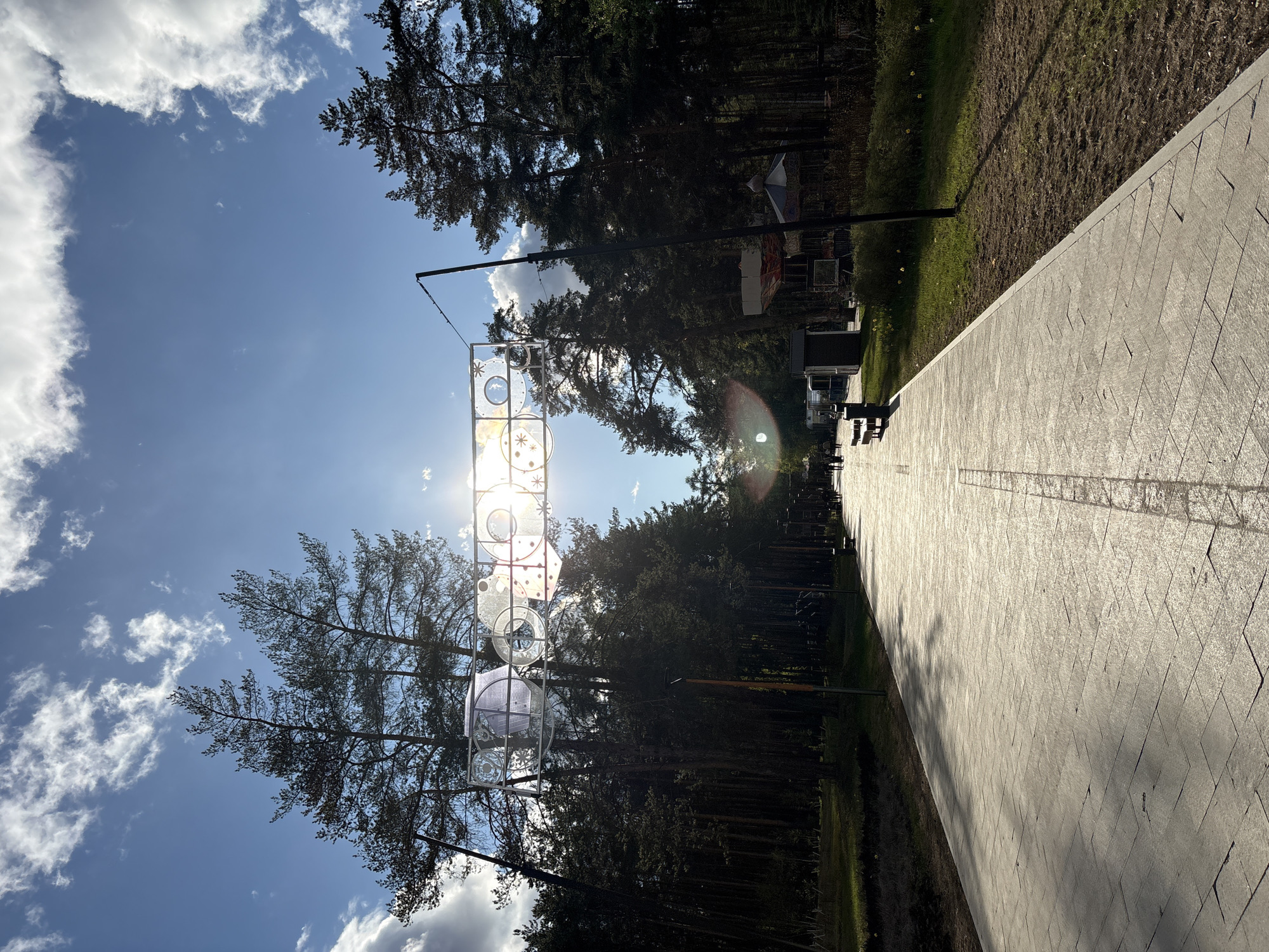 The image shows a pedestrian walkway in the city of Perm, Russia. The walkway is paved with rectangular blocks, and there is a grassy area on one side. Tall trees line the walkway. An art installation made of geometric shapes hangs overhead, partially obscuring the sun. The sky is blue with scattered clouds. There are people sitting on benches along the walkway, but they are distant and indistinct.