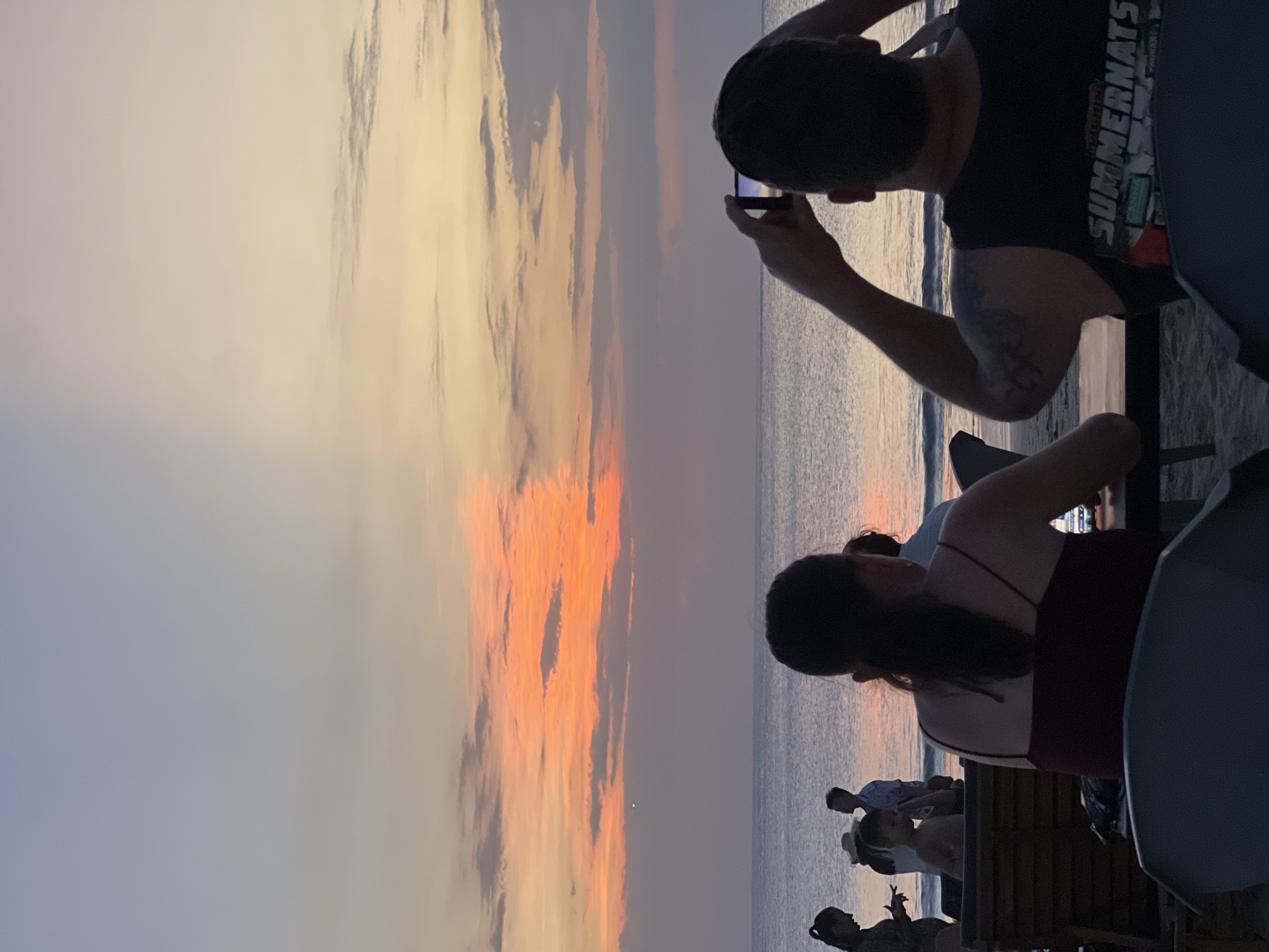 The image captures a sunset scene on a beach in Kuta Selatan, Indonesia. In the foreground, two individuals are seated at tables, their backs to the camera, facing the ocean. One person is holding up a phone to capture the sunset. To the left, a group of people are visible on the beach, including a woman in a hat and a child. The ocean is calm, with gentle waves lapping the shore. The sky is a canvas of warm colors, with hues of orange, pink, and purple dominating the sunset. The atmosphere is serene and relaxed, suggesting a leisurely evening on the beach.