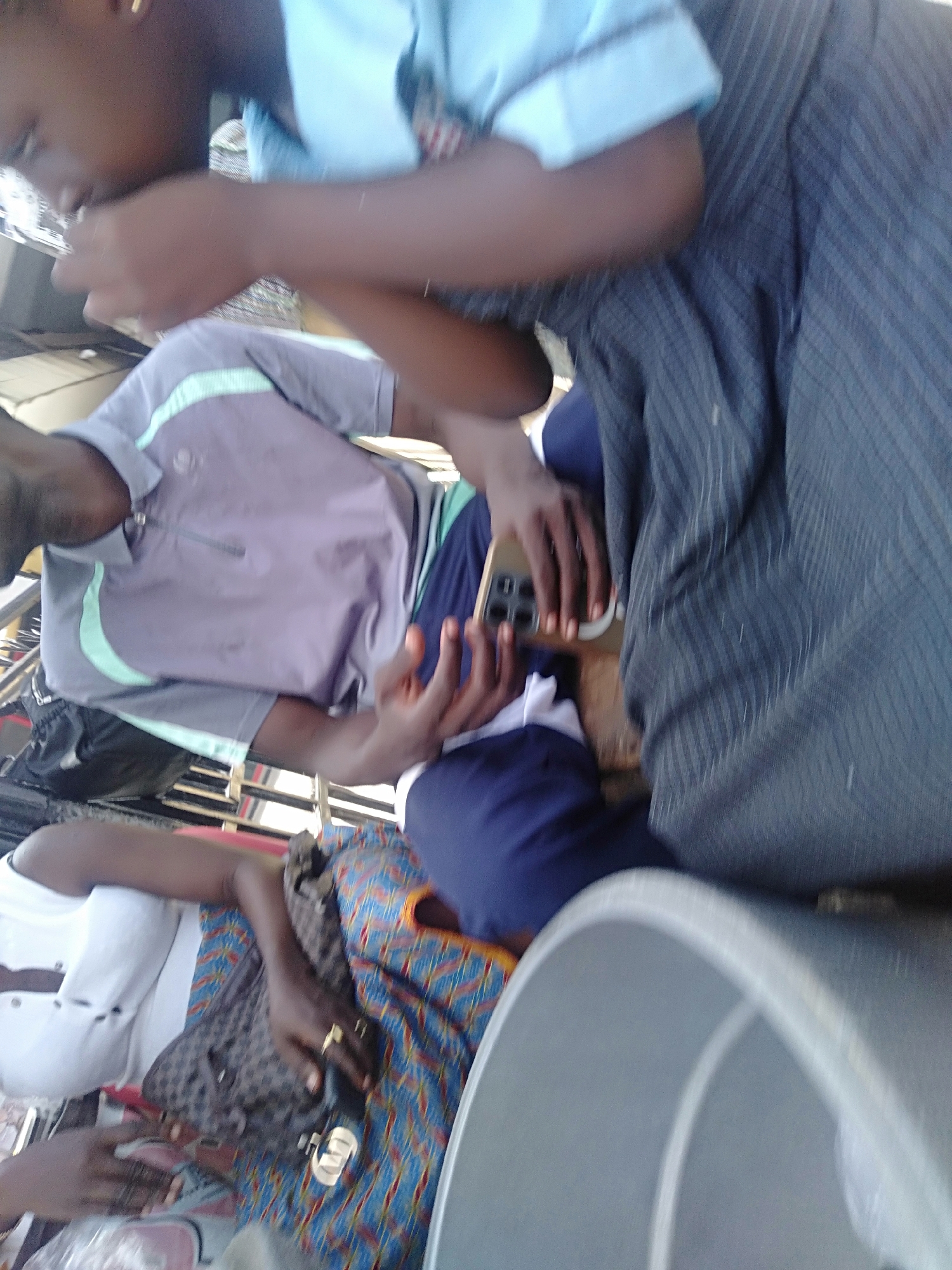 Here is a description of the image:
The image appears to be taken inside a bus or public transportation vehicle, based on the presence of multiple seated individuals and the interior elements. The subjects include people dressed in casual attire, and what appears to be a school uniform. One person is holding a smartphone. Another person is wearing rings. The environment suggests an urban setting. The city, or country, is unknown.
